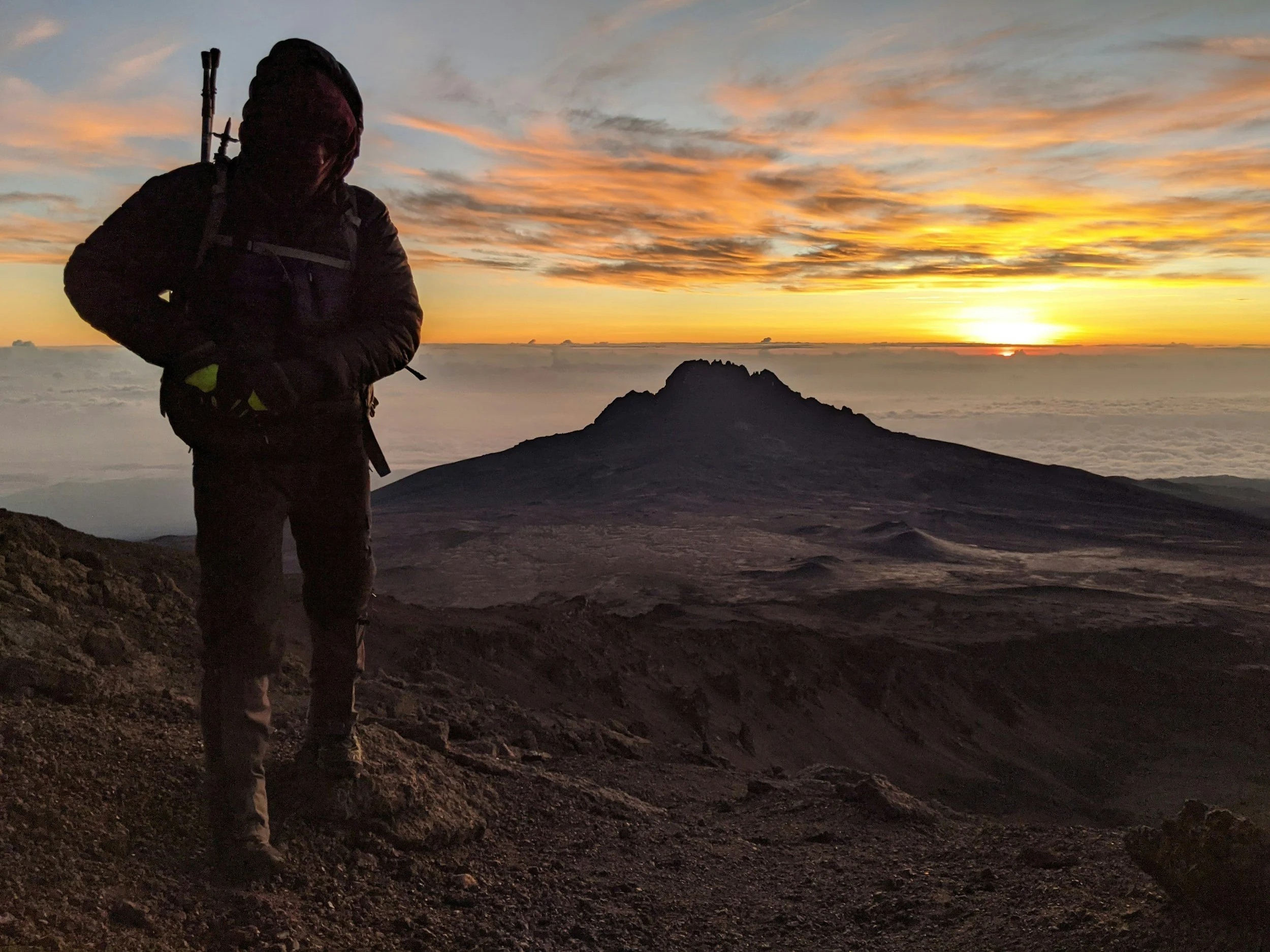 A hiker dressed in dark outdoor gear standing on a rocky mountain slope at sunrise, with a volcano or mountain peak in the background and clouds below on Mount Kilimanjaro.