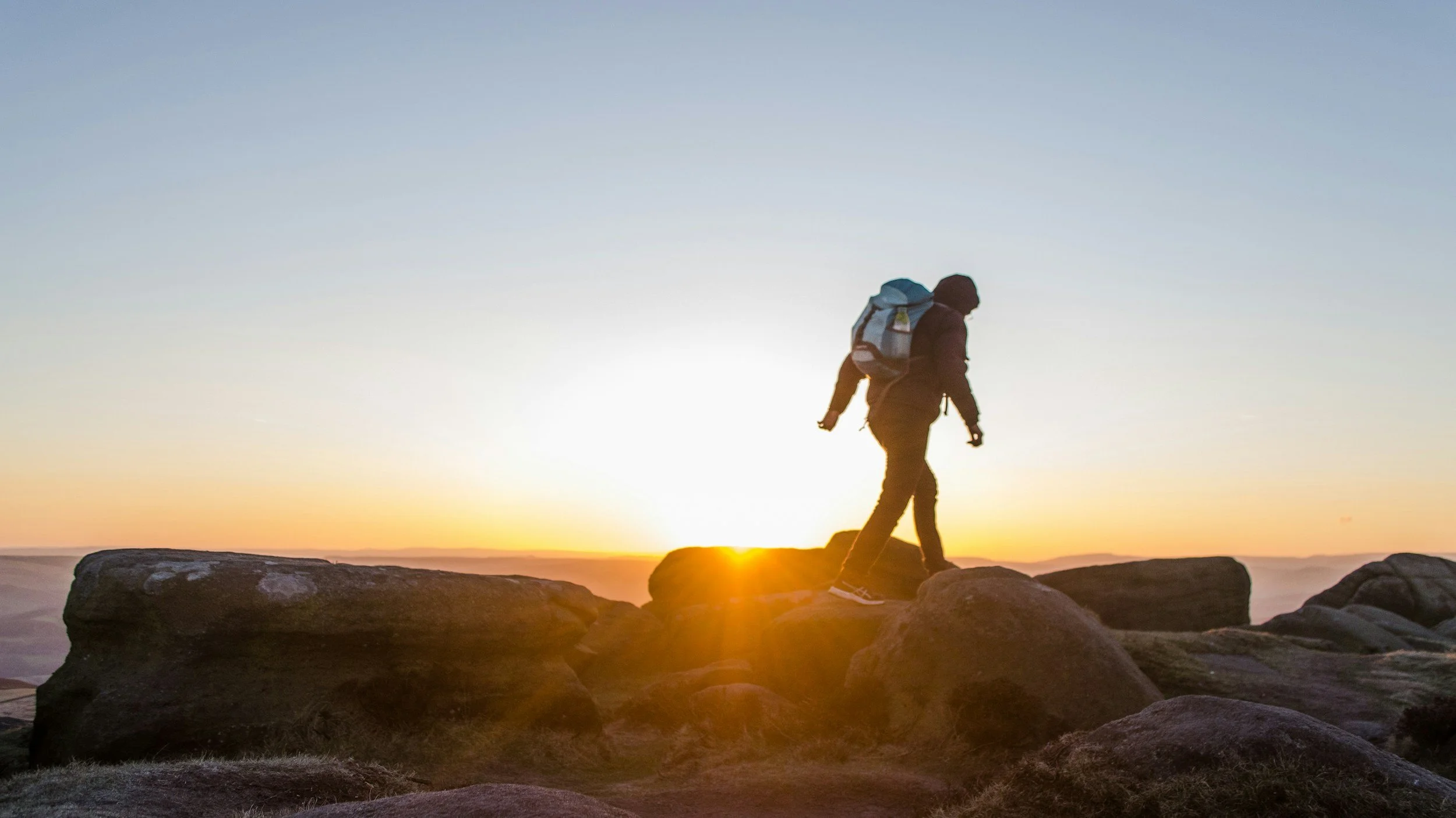 A person hiking with a backpack on rocks during sunrise or sunset, with the sun low on the horizon and a clear sky.