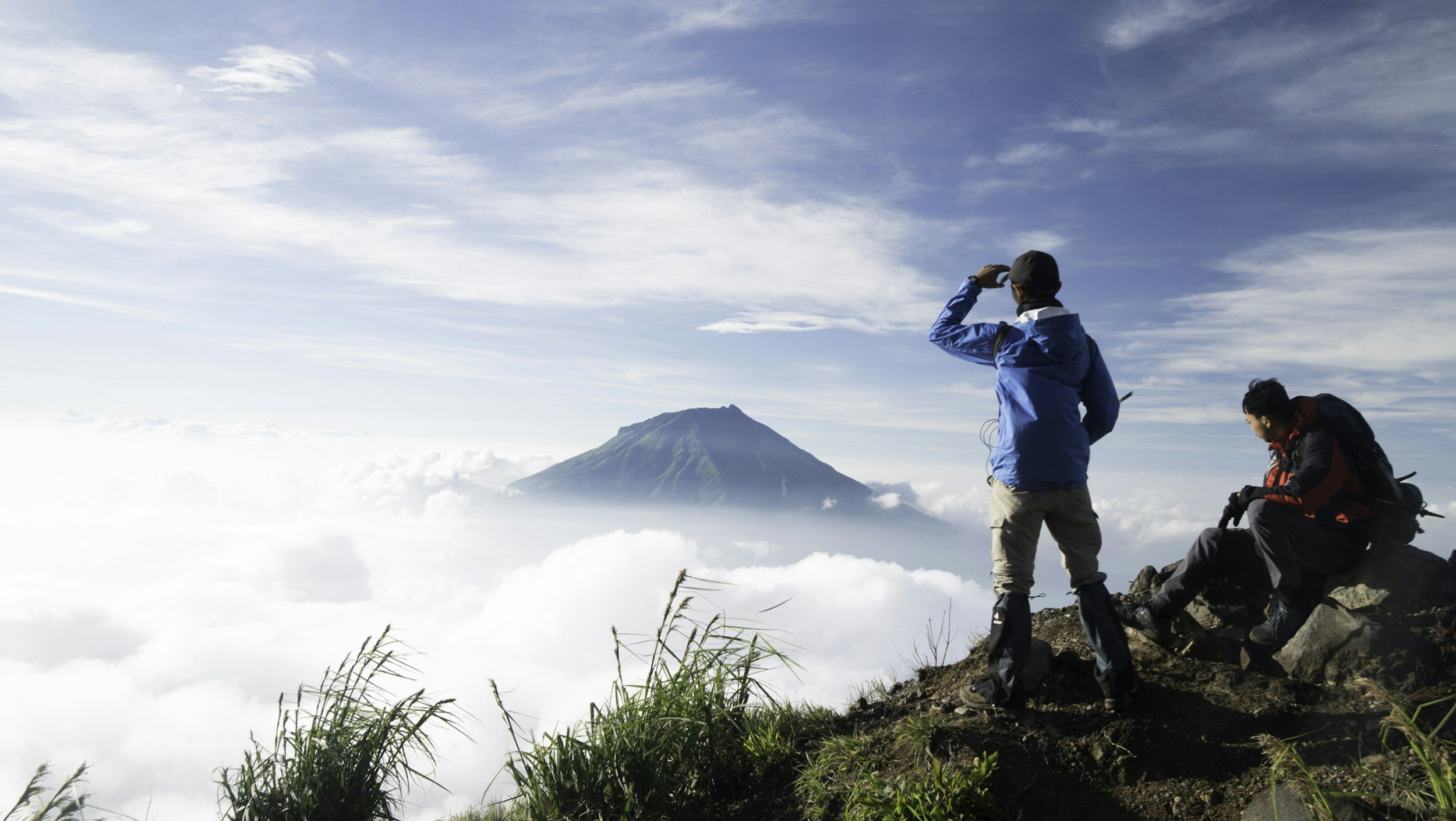 Two hikers on a mountain trail looking at a distant volcano while clouds surround the mountain landscape.  The trekkers are on Mount Kilimanjaro and looking at Mount Meru in Tanzania.