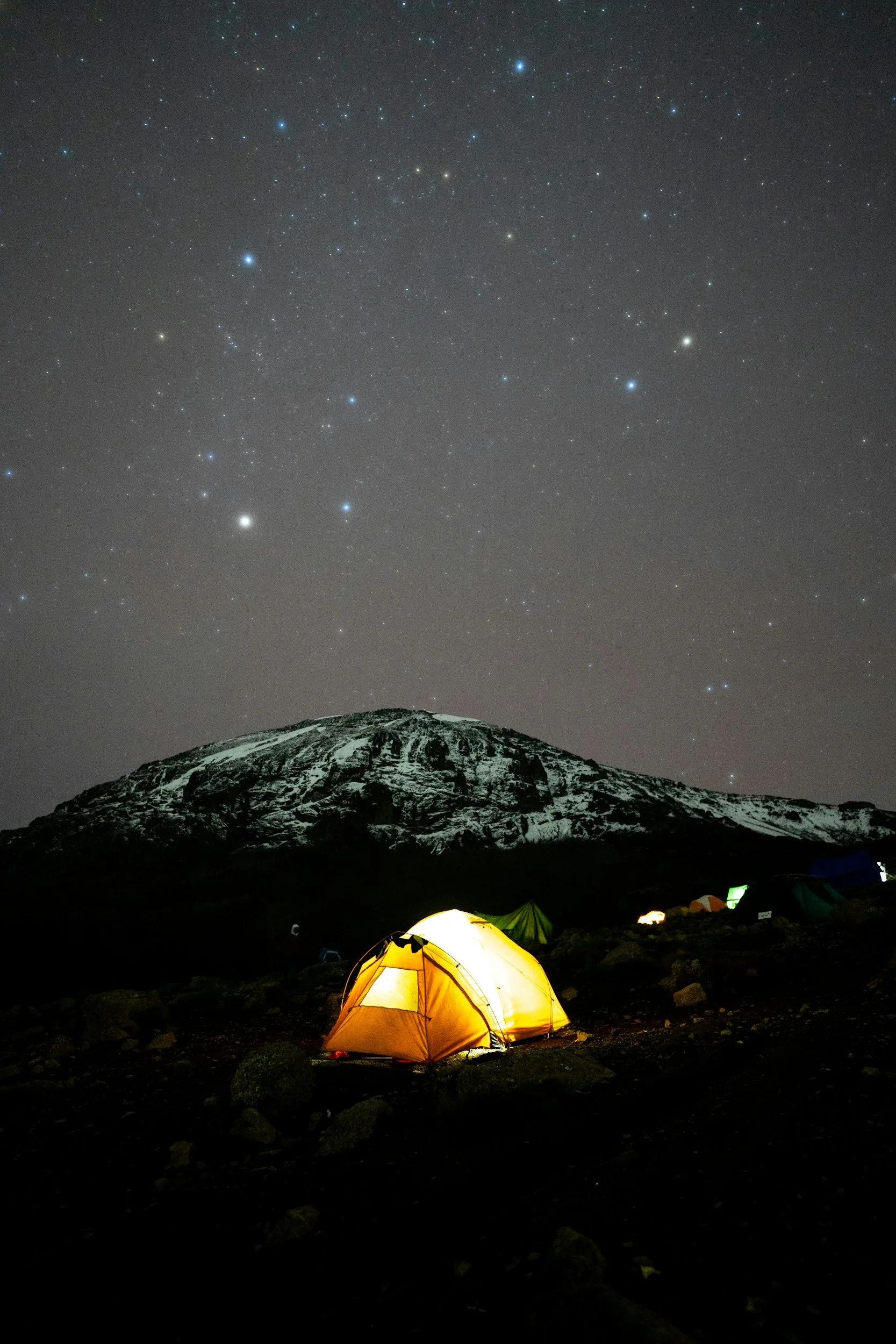 An illuminated yellow camping tent set up on dark terrain with a snow-capped mountain in the background under a star-filled night sky.