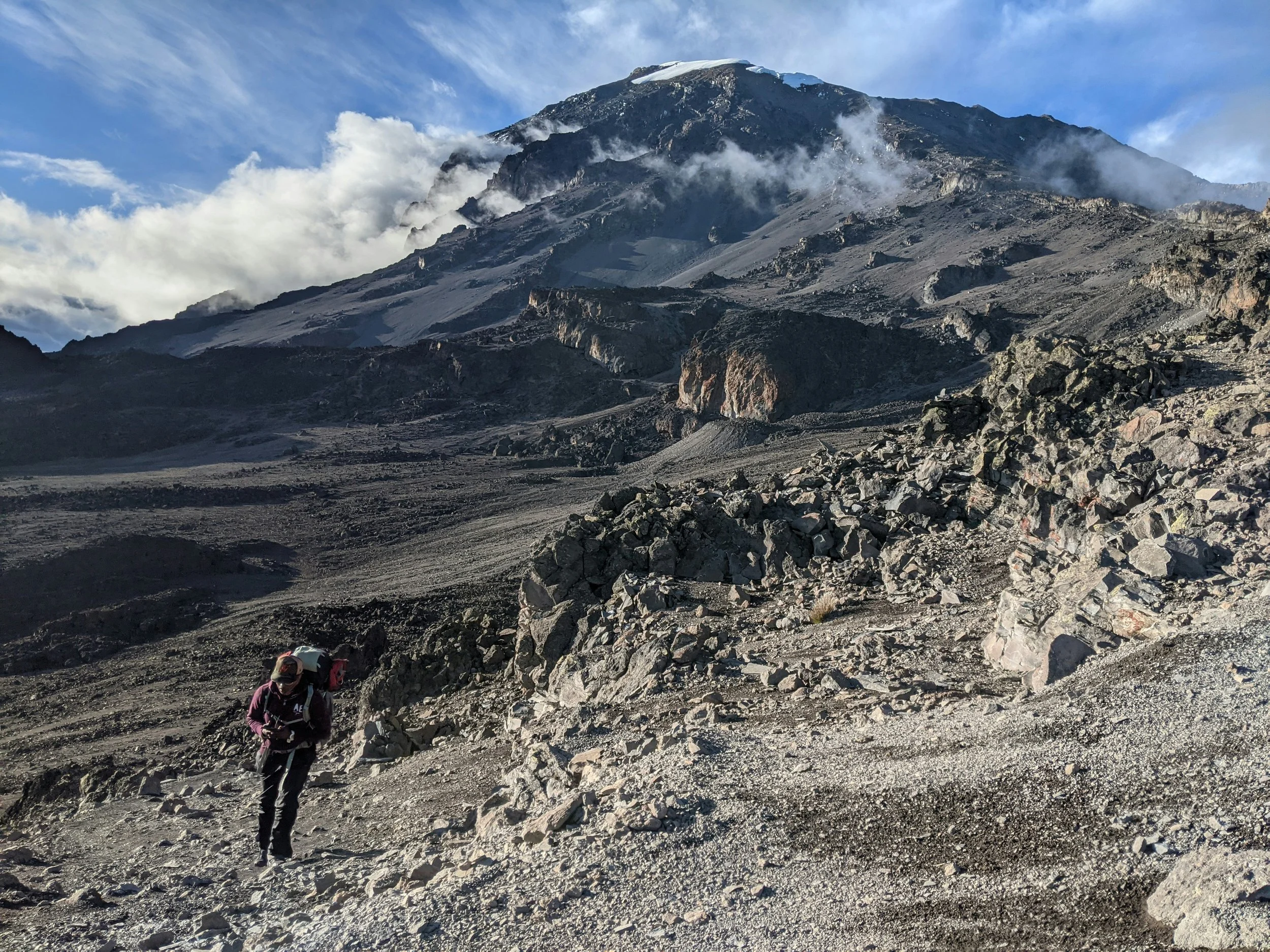 A hiker or porter or guide with a backpack walking on a rocky mountain trail with a large mountain and clouds in the background. Mount Kilimanjaro or Mount Everest Base Camp