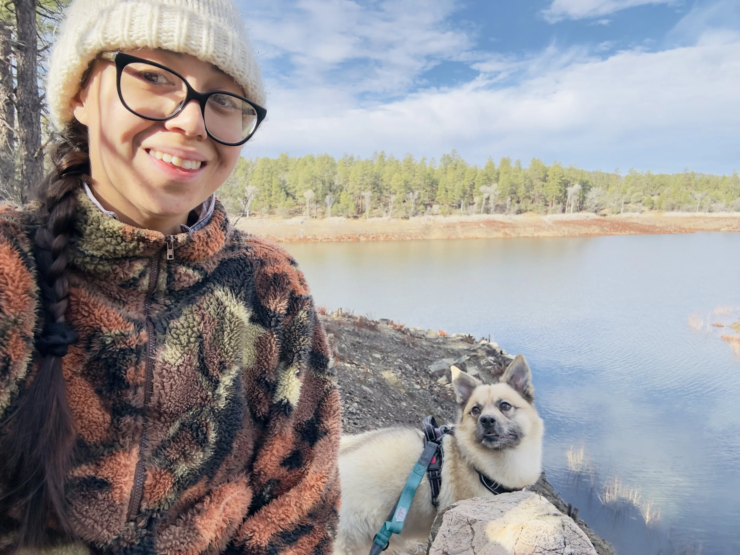 A woman with glasses, wearing a knit hat and a multicolored fleece jacket, smiling outdoors with a lake and forest in the background, accompanied by a small dog on a leash.