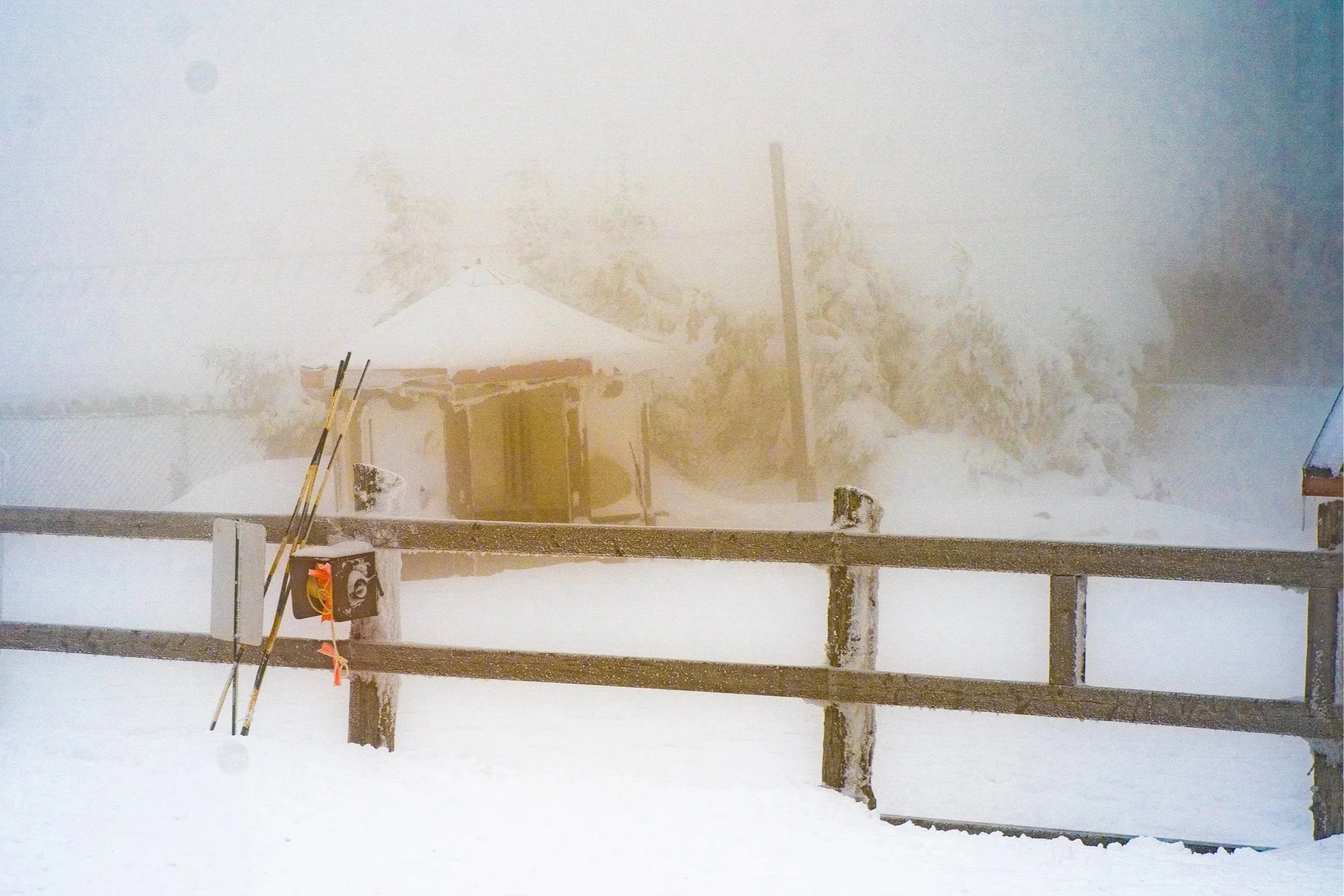 Snow-covered ski slope with a wooden fence and ski poles in the foreground, a small cabin with a pitched roof in the background, and snow-covered trees and fog creating a cold, wintry scene.
