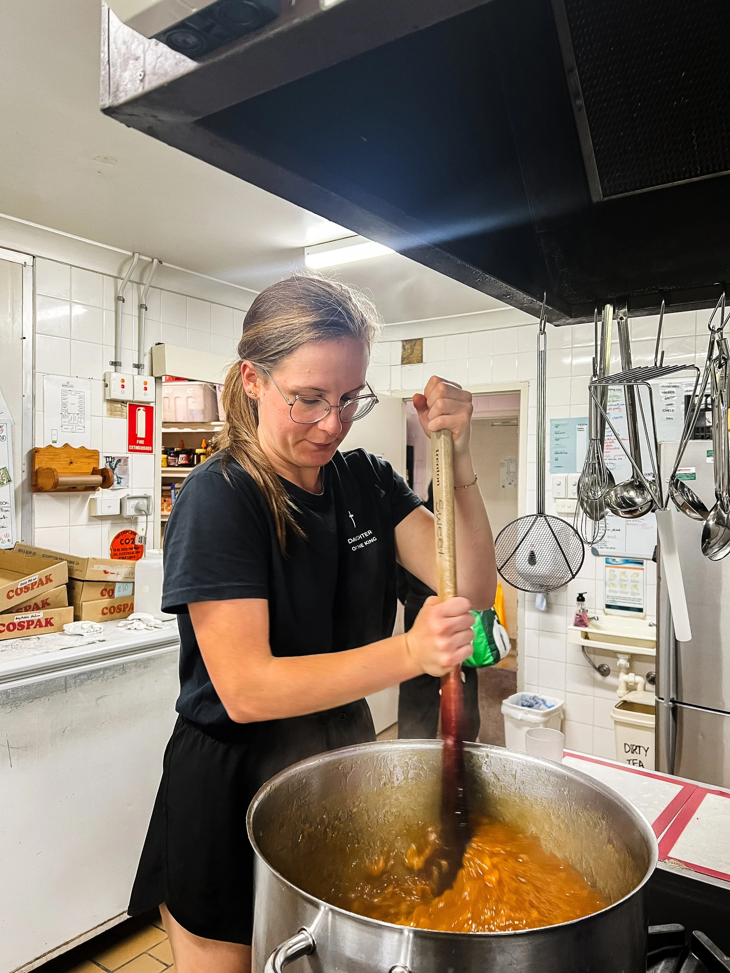 A woman in a kitchen stirring a large pot of orange-colored food with a wooden spoon.