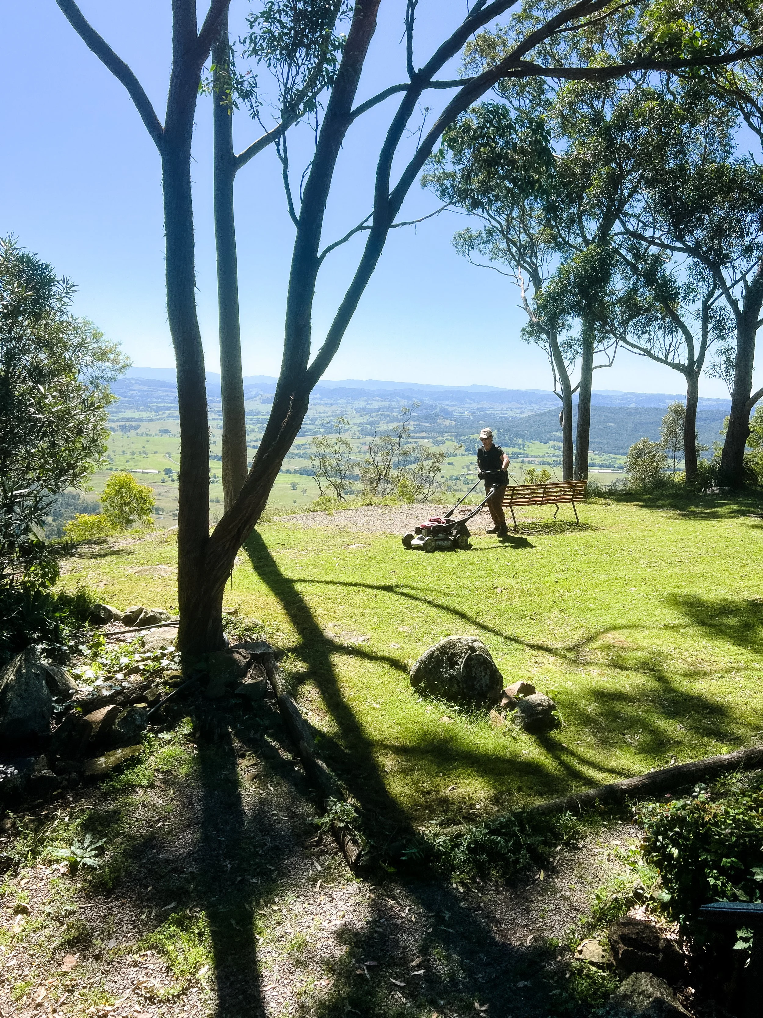 Person mowing the lawn on a lush grassy hilltop with trees, a bench, and scenic view of mountains and valleys in the background.