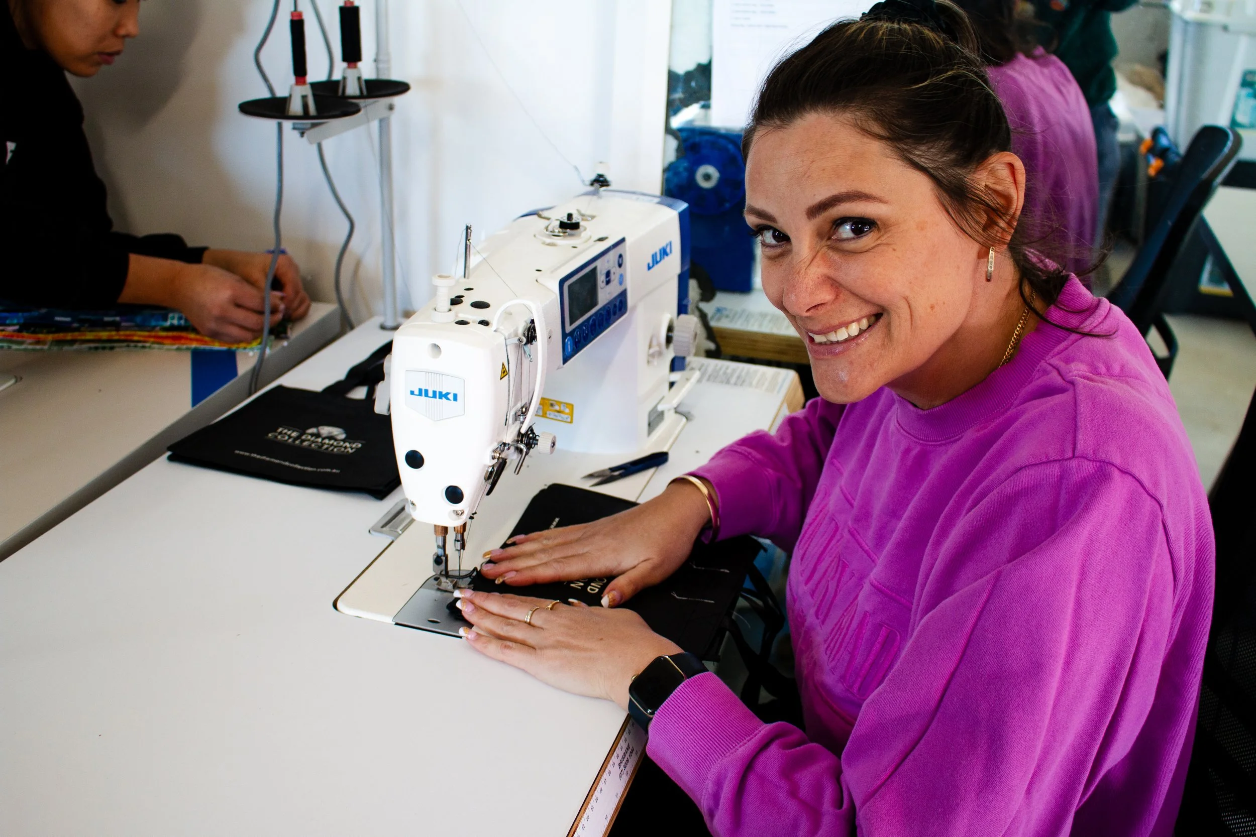 A woman in a pink sweatshirt sewing fabric on a white industrial sewing machine, smiling at the camera. Another person is working on a different sewing machine in the background.