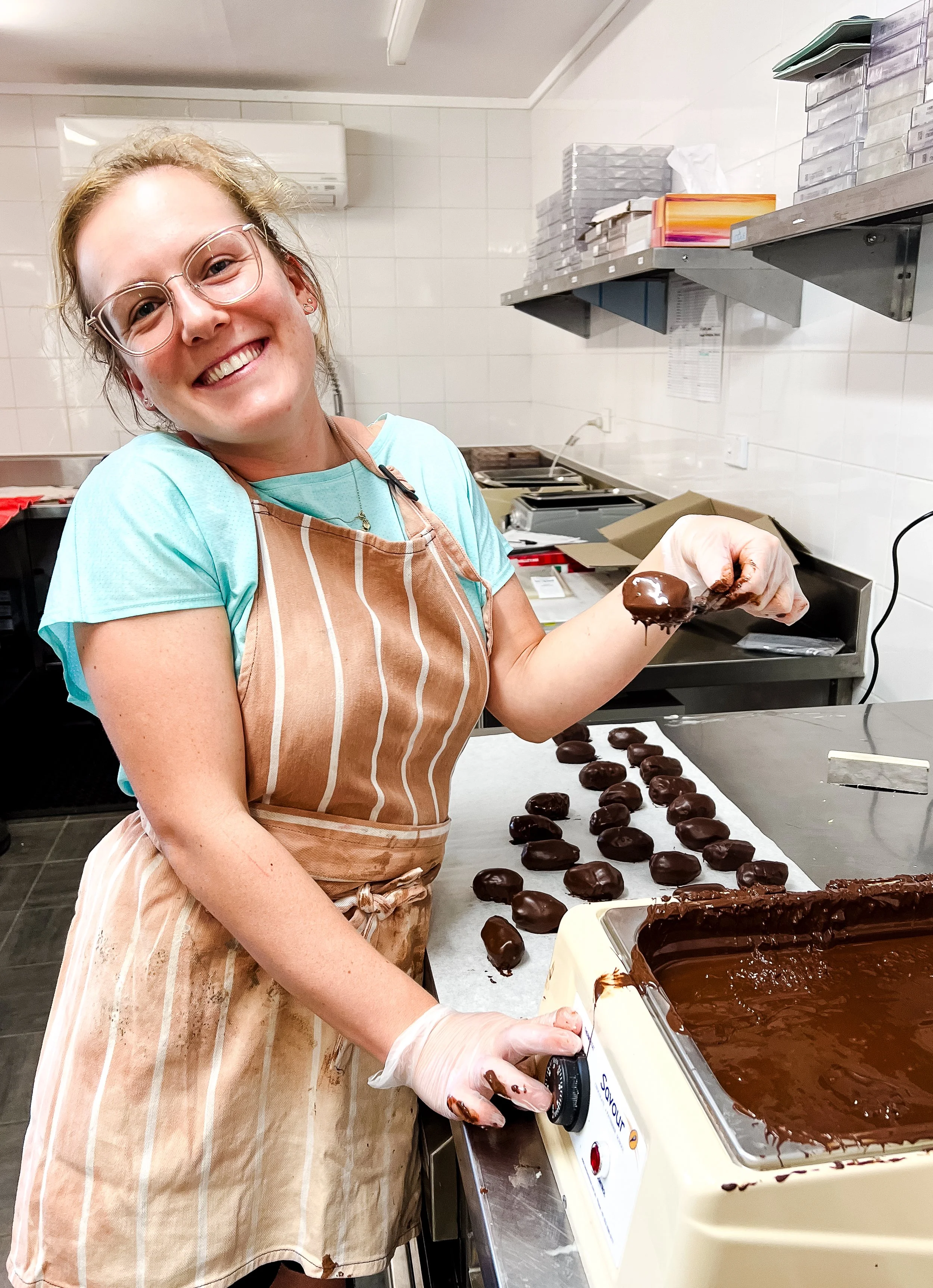 A woman smiling in a bakery or kitchen, holding a chocolate-covered treat, with rows of similar treats on the counter and a large container of melted chocolate nearby.