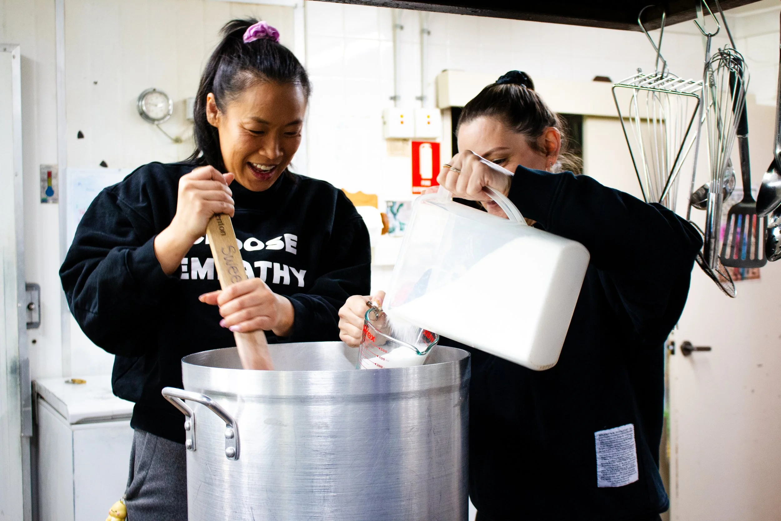 Two women are cooking or baking together, one stirring a large pot while the other pours a bag of flour into it, in a kitchen environment.