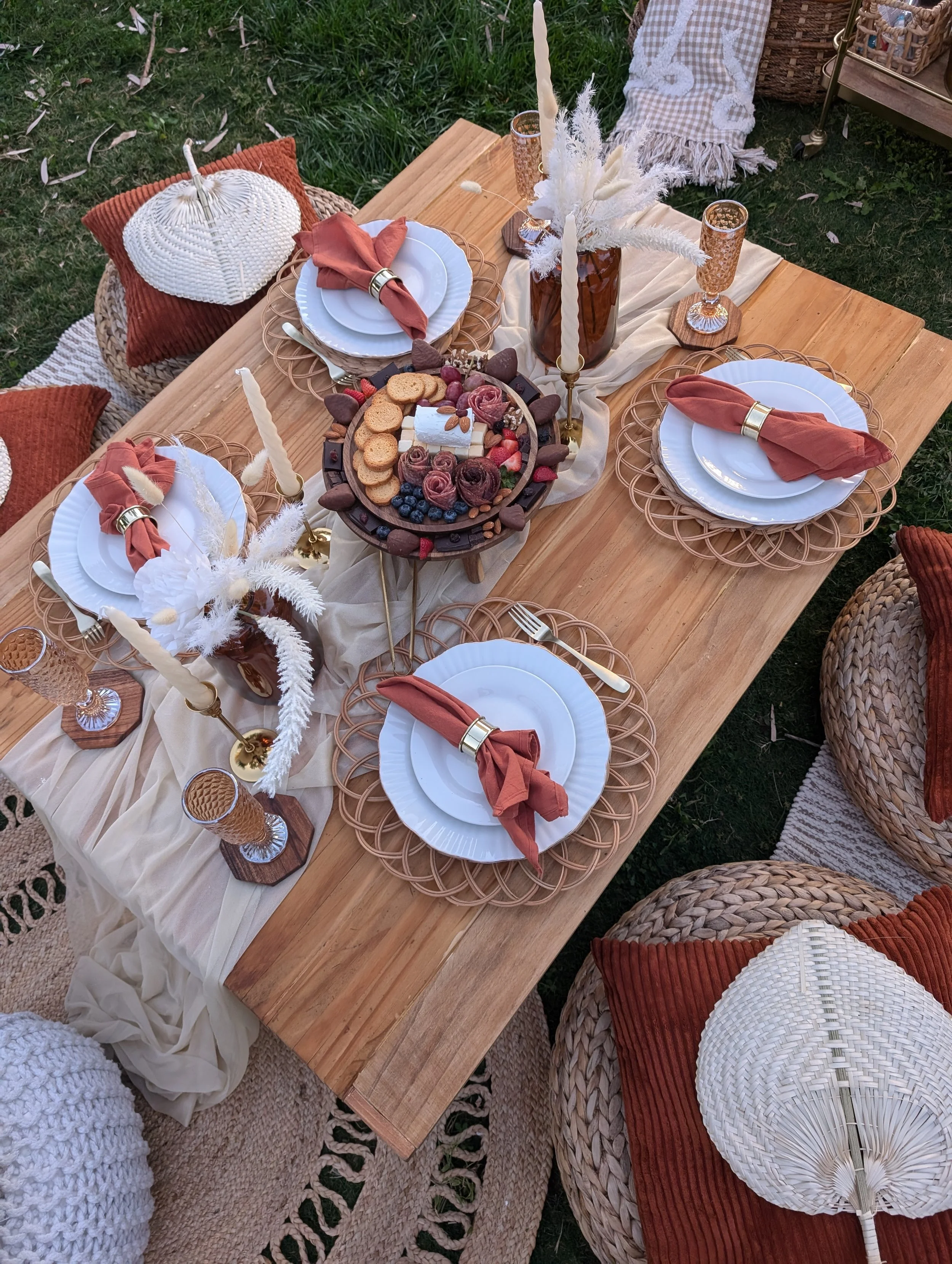 Collection of decorative items including a brass teapot, a stacked brown cloth, a colorful patterned rug, a wooden side table, a yellow and beige patterned rug, a metal lantern, a round orange pouf, and a large beige round rug on a white textured background.