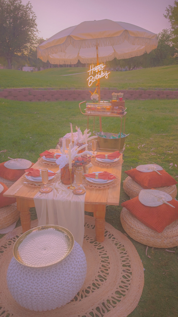 Floor display of home decor items including a brass teapot, colorful patterned rugs, a brown fabric hanging from a table, a wooden side table, a decorative lantern, a yellow and white geometric rug, and a round orange pouf with a white web-like pattern, all against a textured white wall.