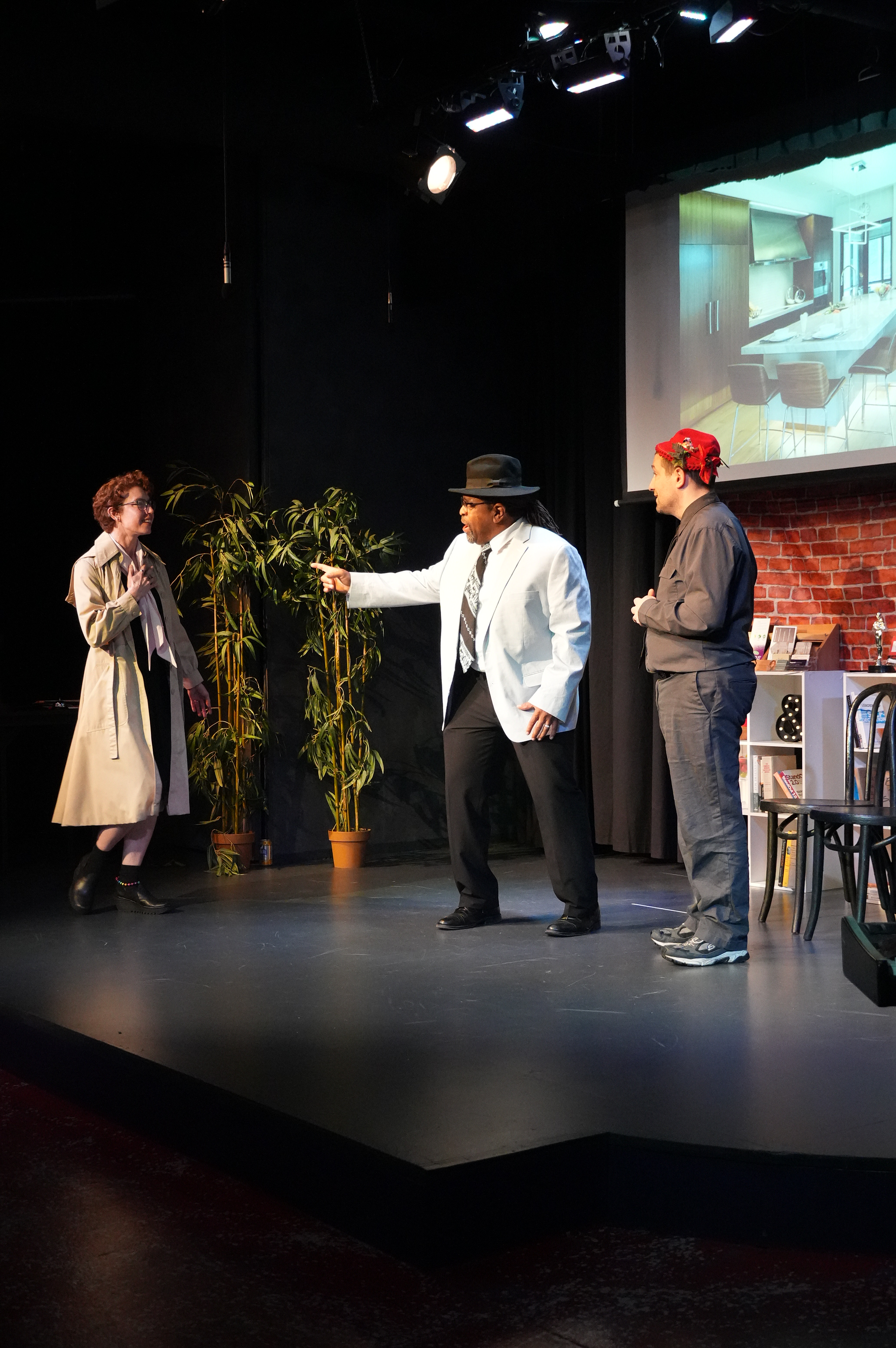 Three actors performing on a stage with a black backdrop, a large plant, and a projected image of a modern kitchen. The actors are engaged in a scene, with one woman in a beige trench coat, a man in a white blazer and a black hat, and another man in