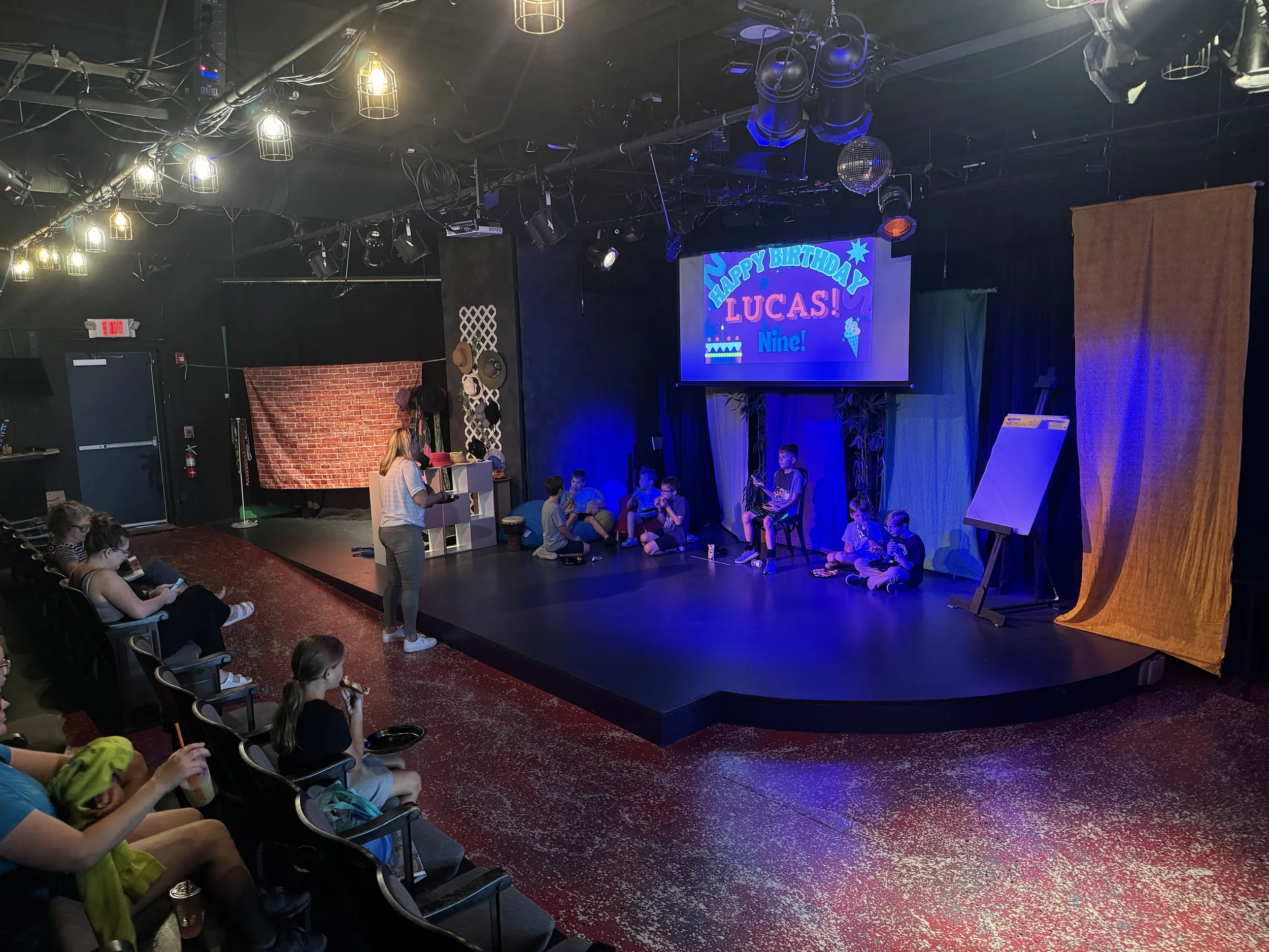 Children performing on a stage with a digital birthday message for Lucas, nine. Audience members watching, some children sitting and some on chairs, in a theater with stage lights and hanging lanterns.