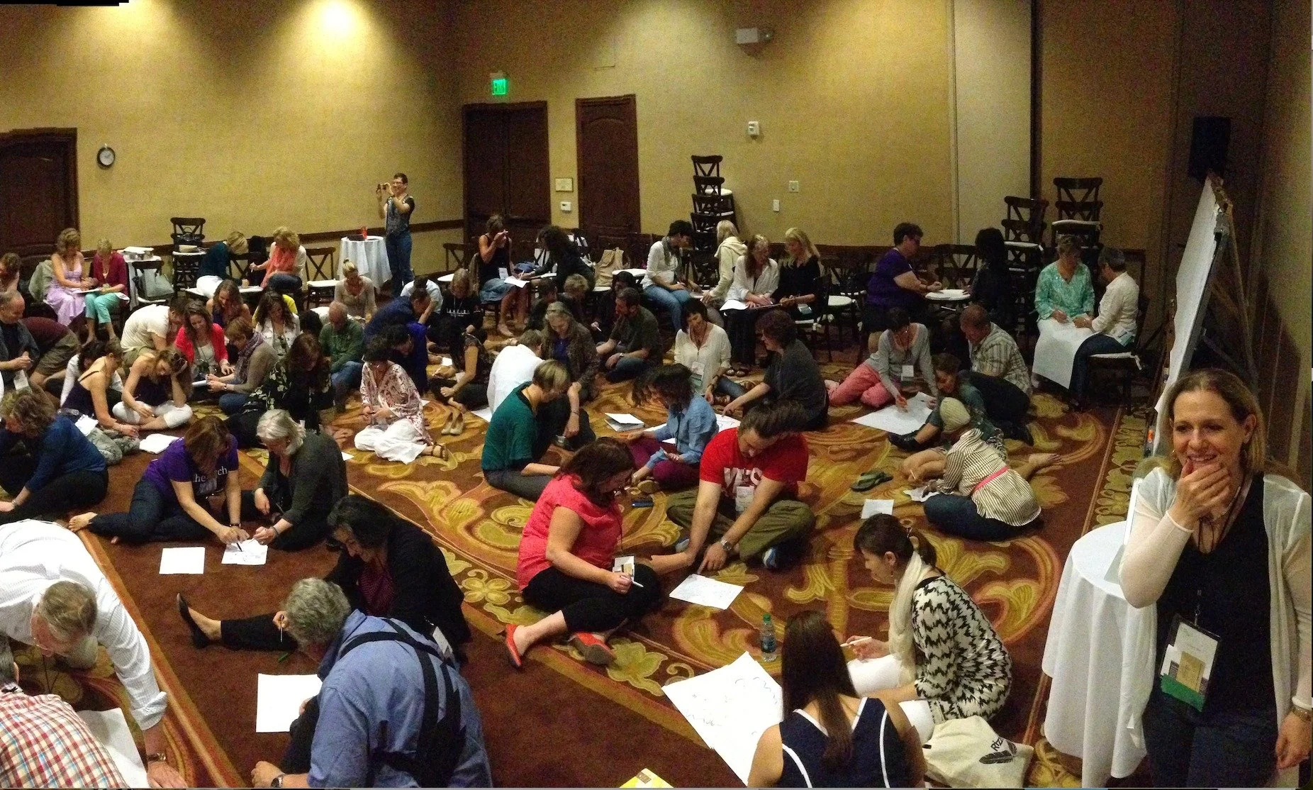 A large group of people engaged in a group activity, sitting on the floor with papers and drawing materials, in a multi-purpose room.