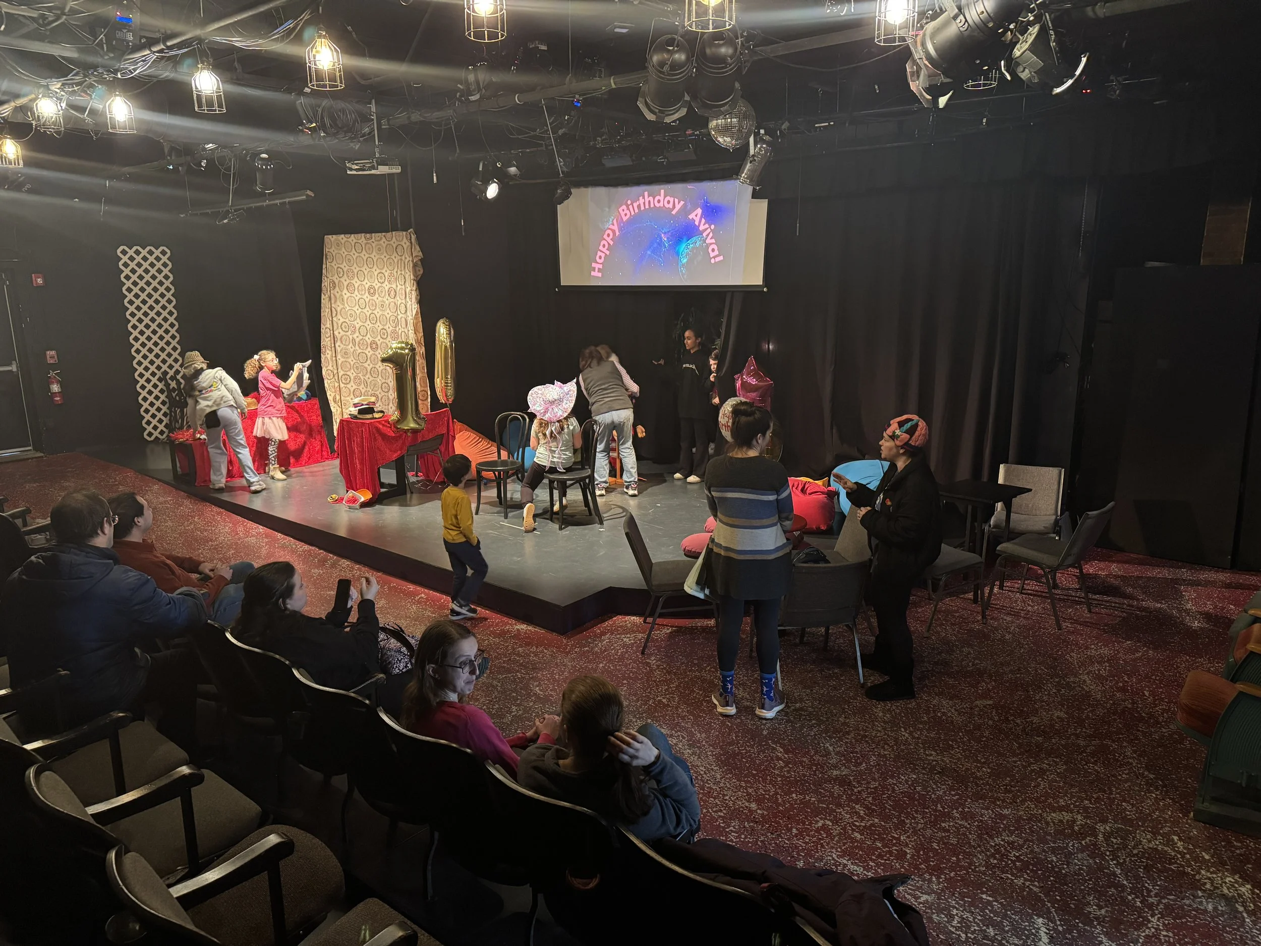 Children and adults preparing for a children's birthday party on a small theater stage with decorations and a large screen displaying a 'Happy Birthday' message.