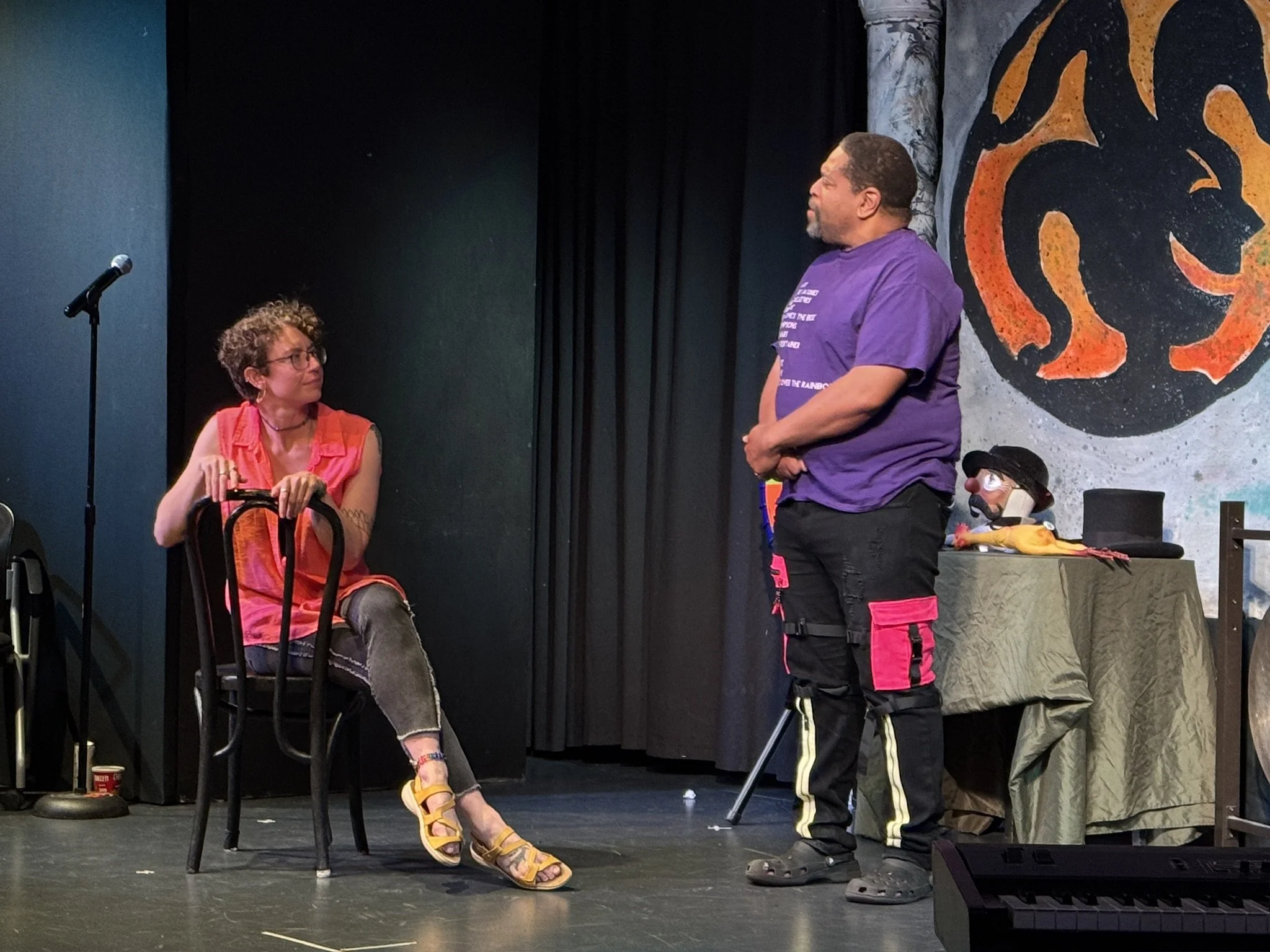 A woman sitting on a black chair on stage, talking to a man standing in front of her, with a microphone stand to the left. There is a table with theatrical props, including a puppet wearing a hat, behind them. A large emblem of a lion's head is paint