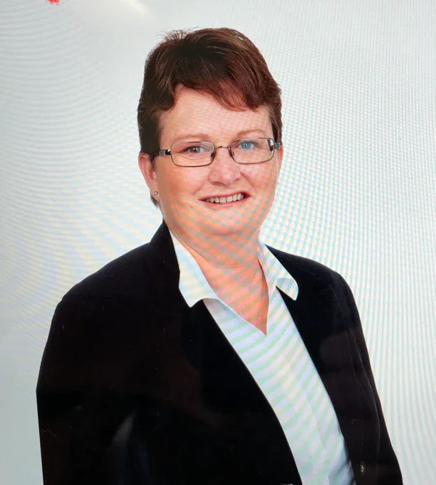 A middle-aged woman with short brown hair, glasses, wearing a black blazer and a white shirt, smiling at the camera against a plain light background.