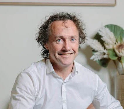 A man with curly hair smiling, wearing a white shirt, sitting indoors with a flower arrangement in the background.