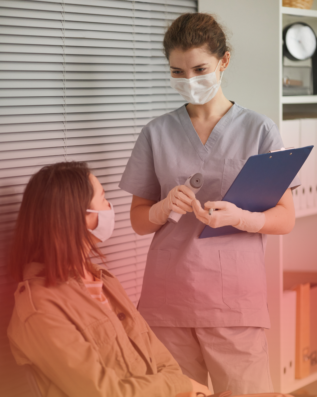 A nurse in scrubs and a face mask holding a clipboard and a medical instrument, standing next to a seated patient with a face mask, in a medical setting.