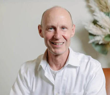 A smiling middle-aged man with a shaved head wearing a white button-down shirt, sitting indoors with a white decorative tree in the background.