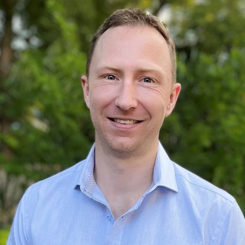 A smiling man with short brown hair wearing a light blue collared shirt outdoors with green foliage in the background.