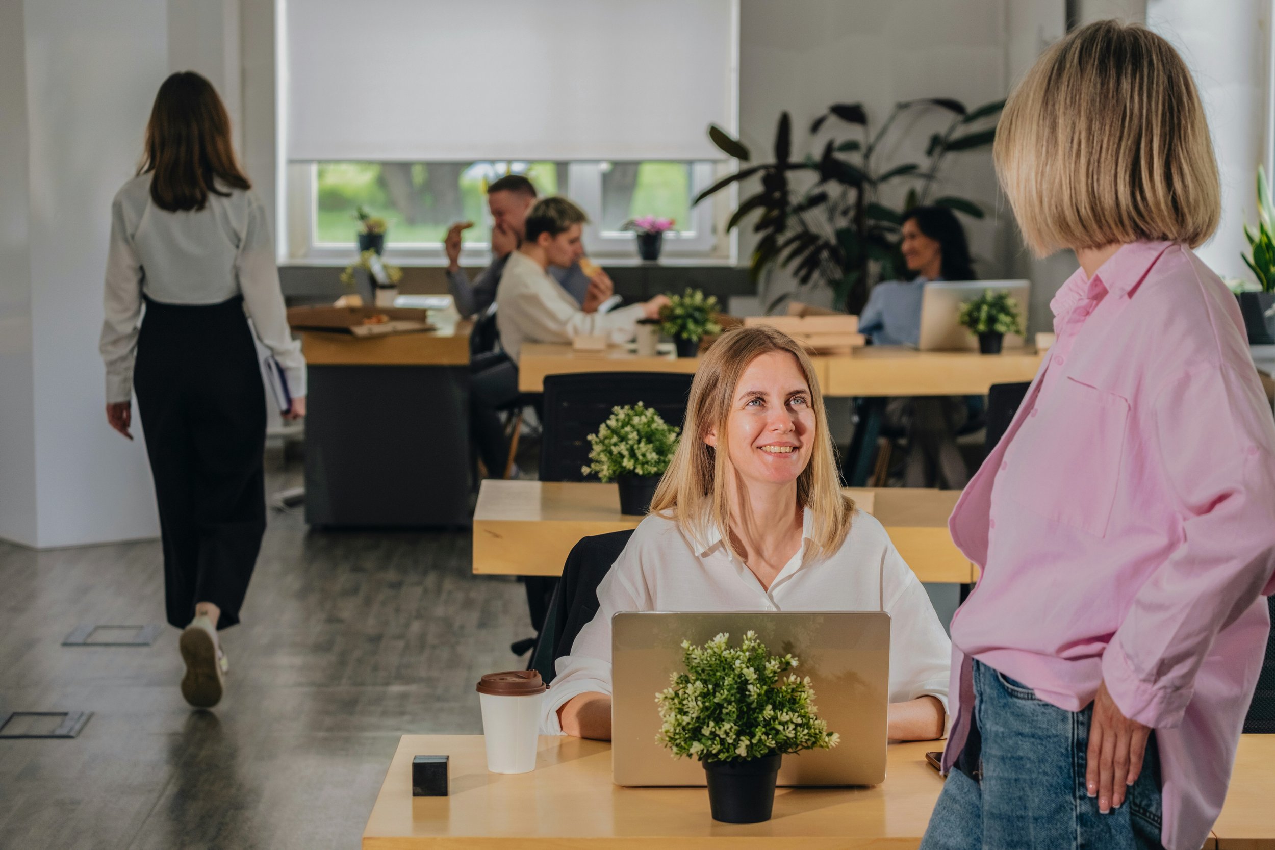A woman sitting at a desk with a laptop, smiling and looking at another woman standing in front of her in a modern office space. The office has wooden tables, potted plants, and large windows.