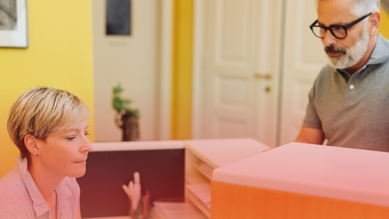 A woman sitting at a desk or table, looking ahead, with a man standing nearby and looking down, in a room with yellow walls.