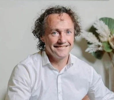 A man with curly hair wearing a white shirt smiling at the camera, sitting in front of a plant and flowers.