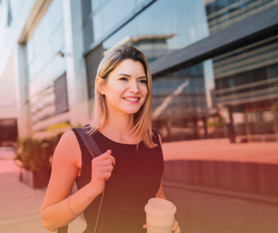 A young woman holding a coffee cup, smiling, outside a modern glass building.