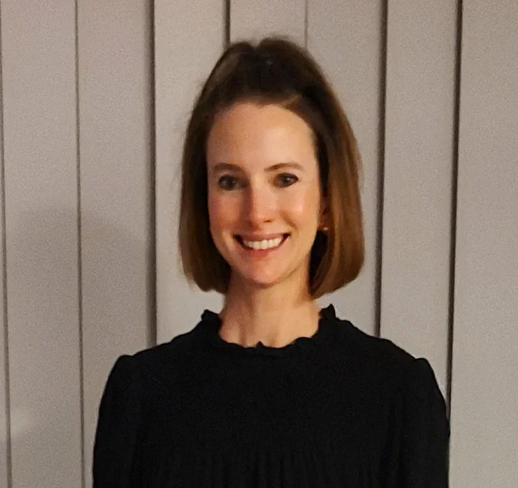 A woman with shoulder-length brown hair smiling at the camera, wearing a black top, standing in front of a light gray wooden wall.