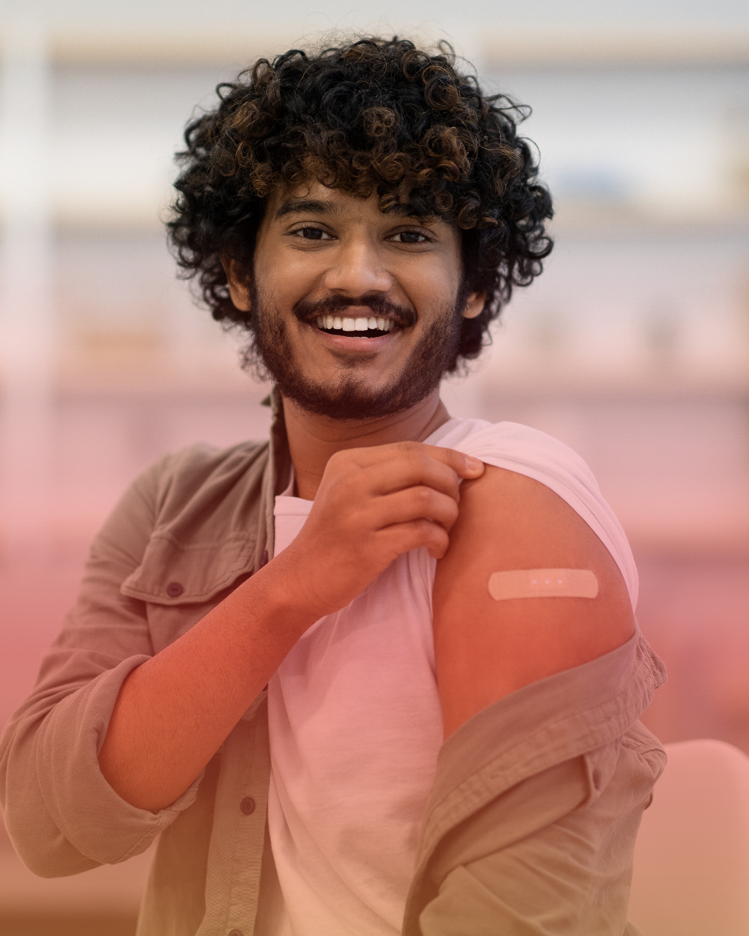 Young man with curly hair and a beard smiling, showing a bandage on his upper arm after vaccination, with his shirt partially off to reveal the bandage.