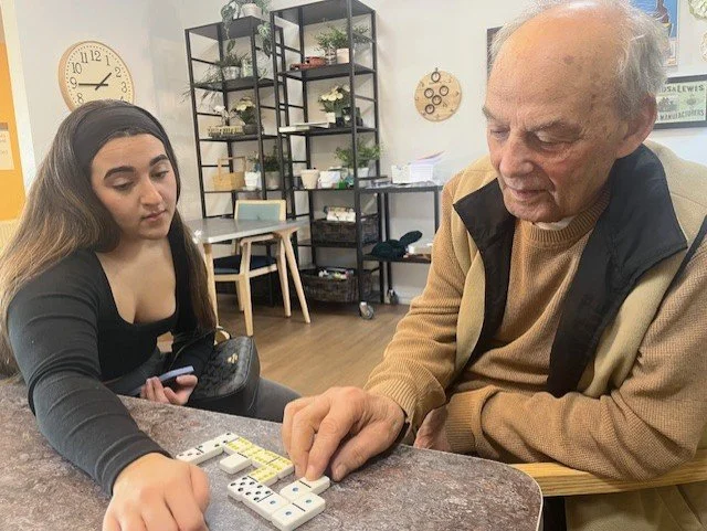 An elderly man and a young woman are sitting at a table playing dominoes. The man is touching the dominoes, and the woman is observing. The background features a clock, shelves with plants, and some decor on the wall.