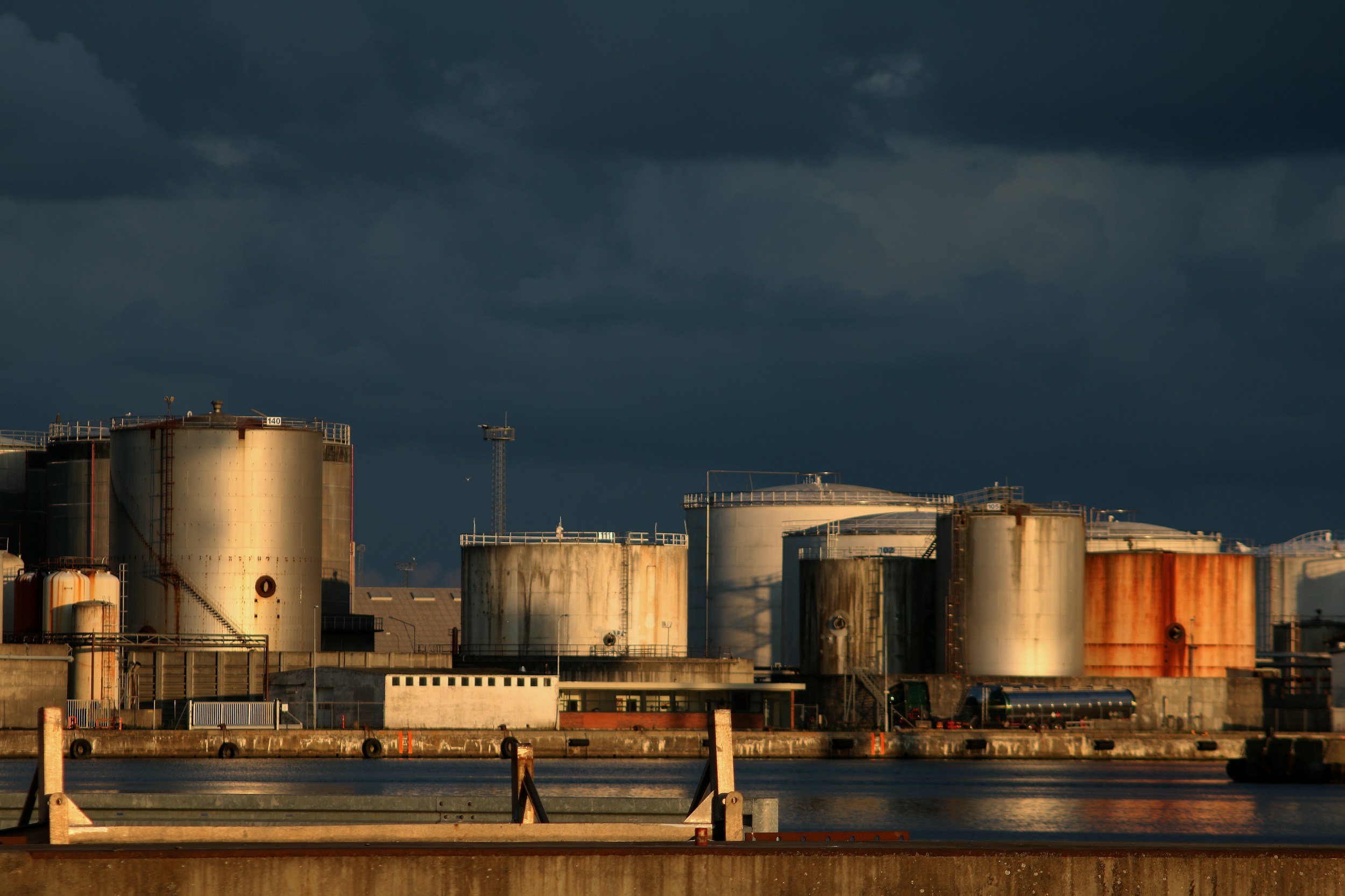 Industrial storage tanks along a waterfront under dark, cloudy sky.