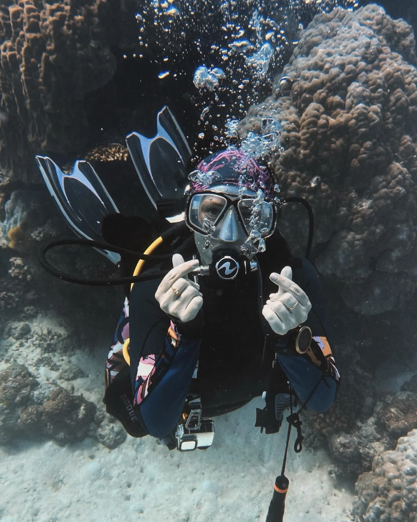A scuba diver underwater among rocks, holding a snorkel mask with both hands, with bubbles rising from the mask.