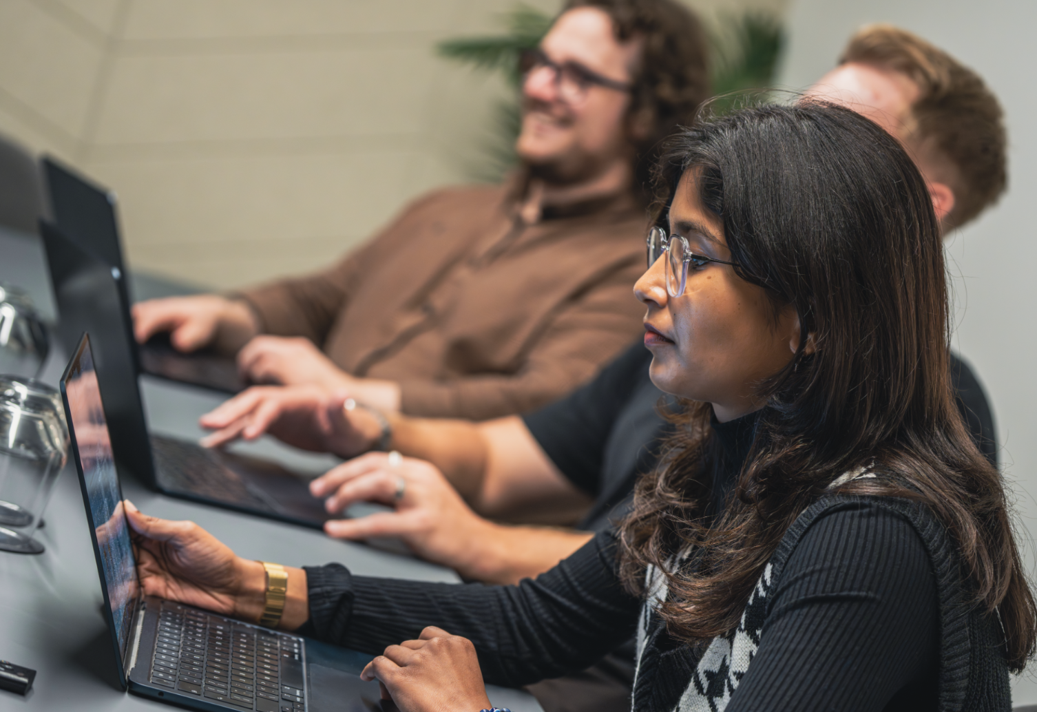 Three people sitting at a conference table working on laptops during a meeting or workshop, with a woman in the foreground and two men blurred in the background.