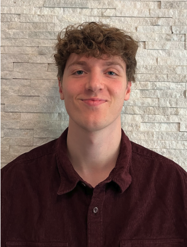 Young man with curly hair smiling, wearing a maroon button-up shirt, standing in front of a textured white brick wall.