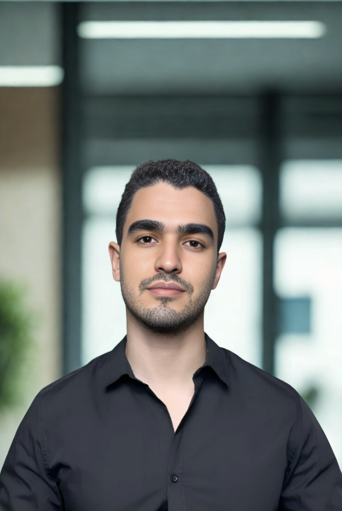 Headshot of a young man with short dark hair, wearing a black collared shirt, standing in front of a blurred office background with windows.