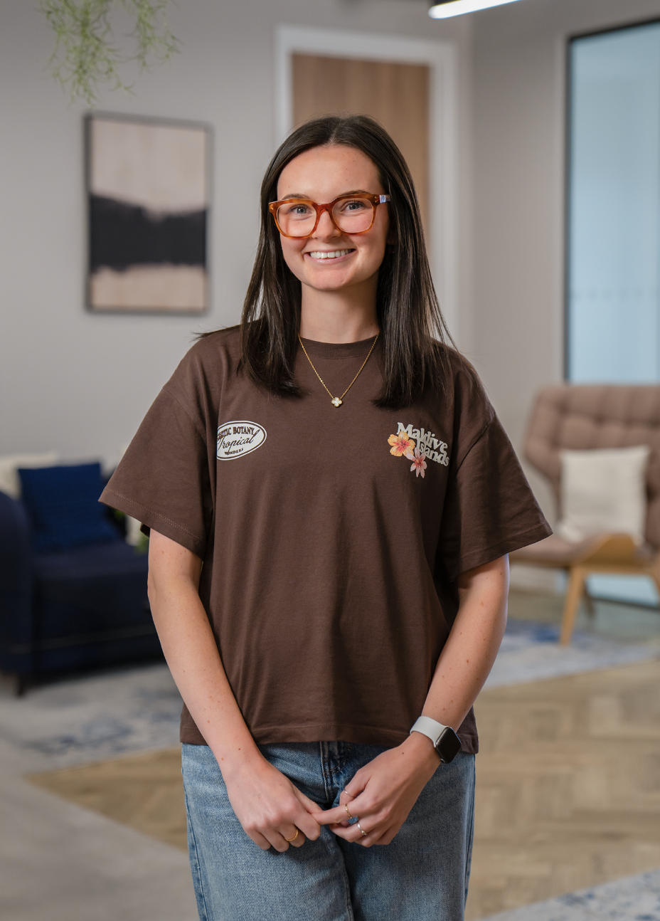 A woman with dark hair, glasses, and a brown T-shirt standing in a modern interior with a background that includes a blue sofa, a plant, and an abstract painting.