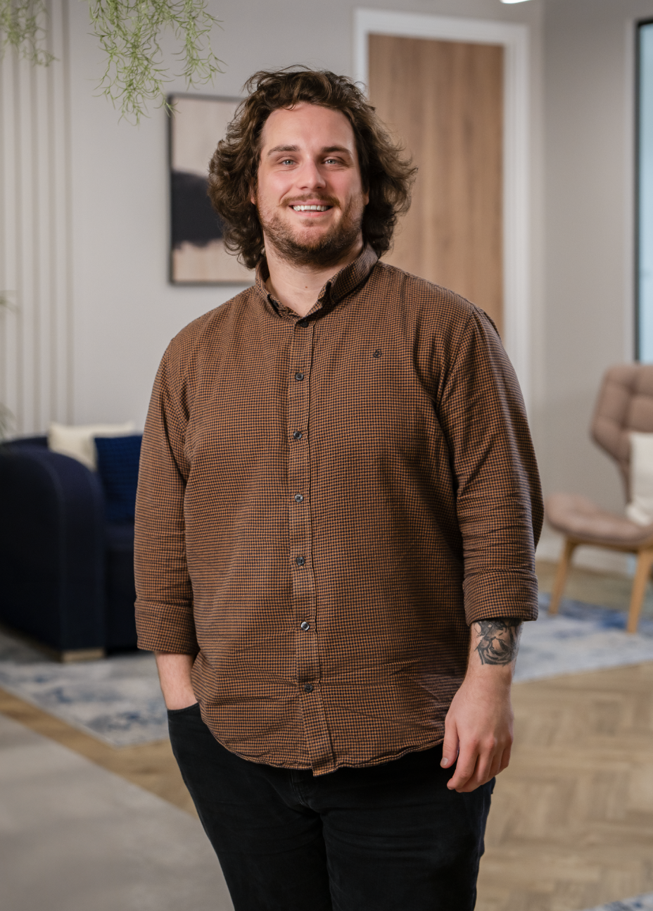 A smiling man with curly brown hair, wearing a brown checkered shirt and black pants, standing in a modern living room.