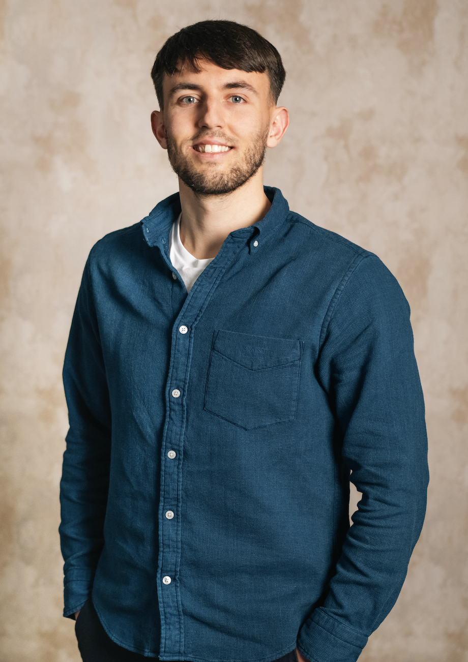 Young man with a beard and short brown hair, wearing a navy blue button-up shirt over a white t-shirt, smiling and standing against a beige textured background.