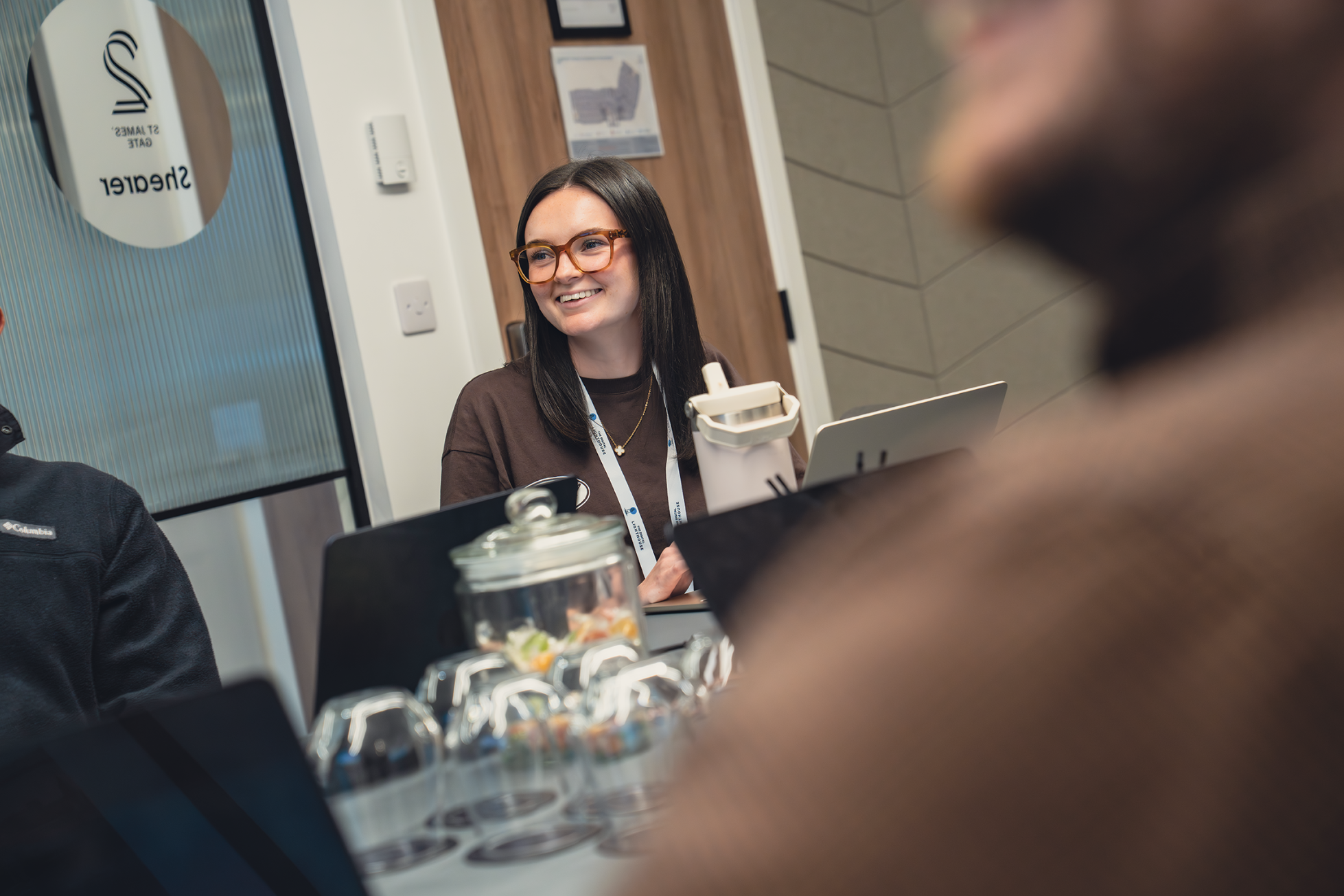 A woman with dark hair and glasses smiling while sitting at a table during a meeting or conference, with a group of people around her.