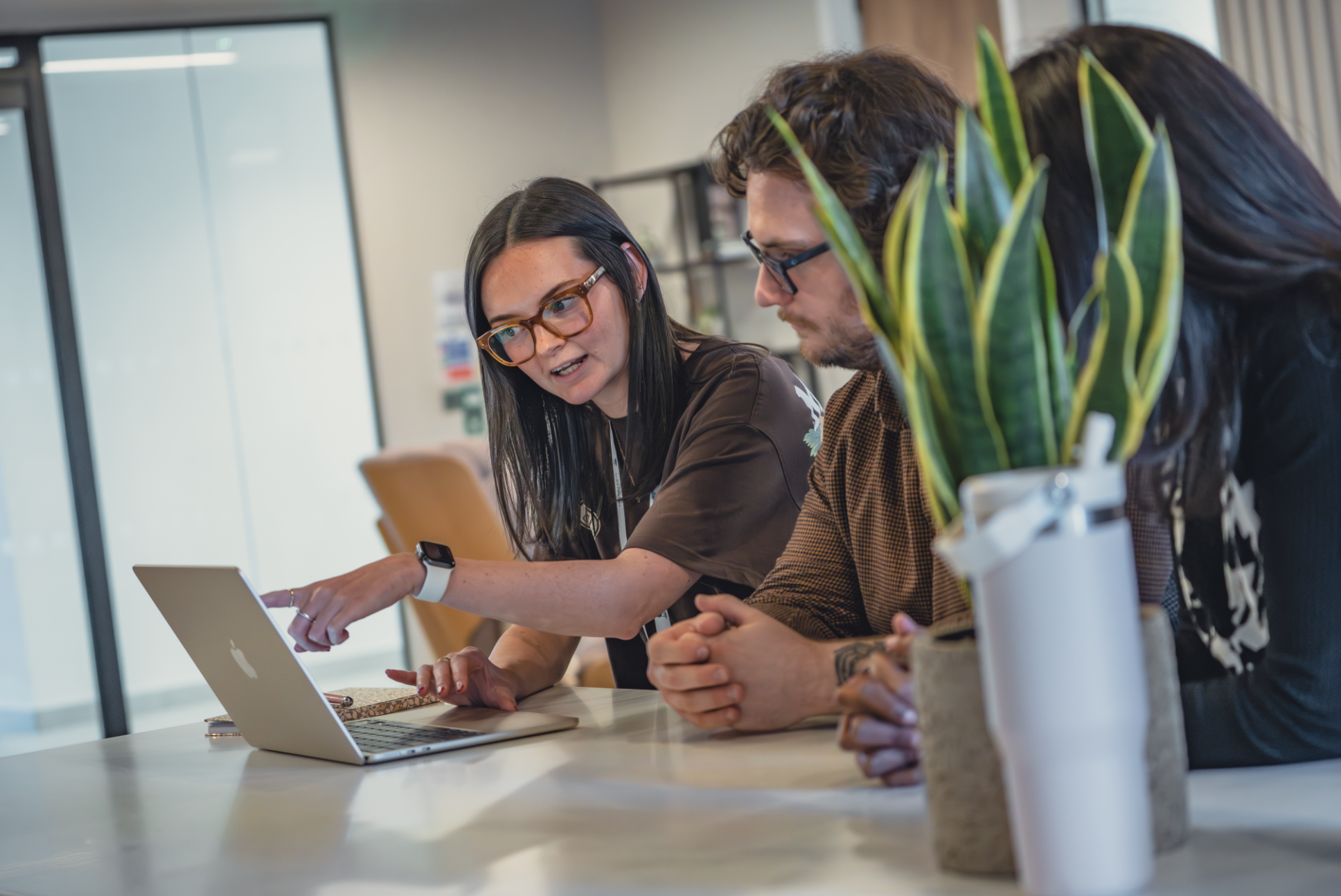 Three people sitting at a conference table engaged in a discussion, with a woman pointing at a laptop screen.
