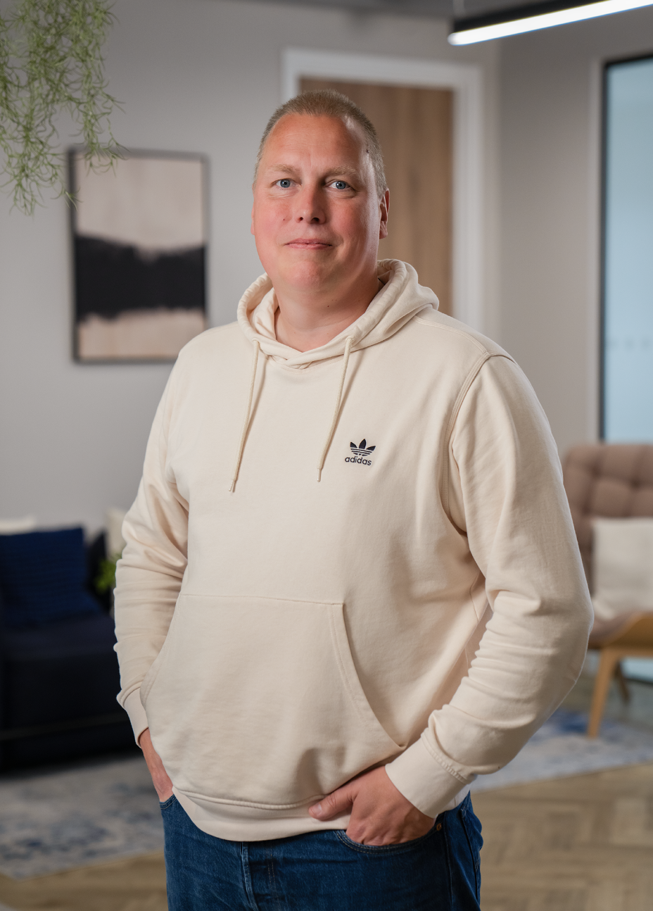 A man with short blonde hair wearing a cream-colored Adidas hoodie and blue jeans standing in a modern living room with decor and a large window.