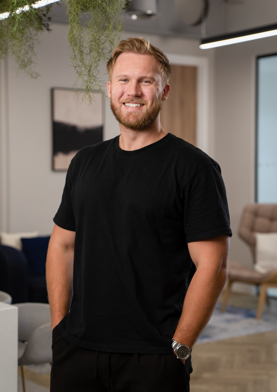 A smiling young man with a beard and short red hair standing indoors in a modern, well-lit room with green plants and contemporary furniture.