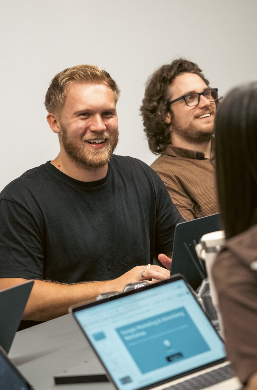 Two men sitting at a table in a meeting room, smiling and engaged in conversation, with laptops and a tablet in front of them.