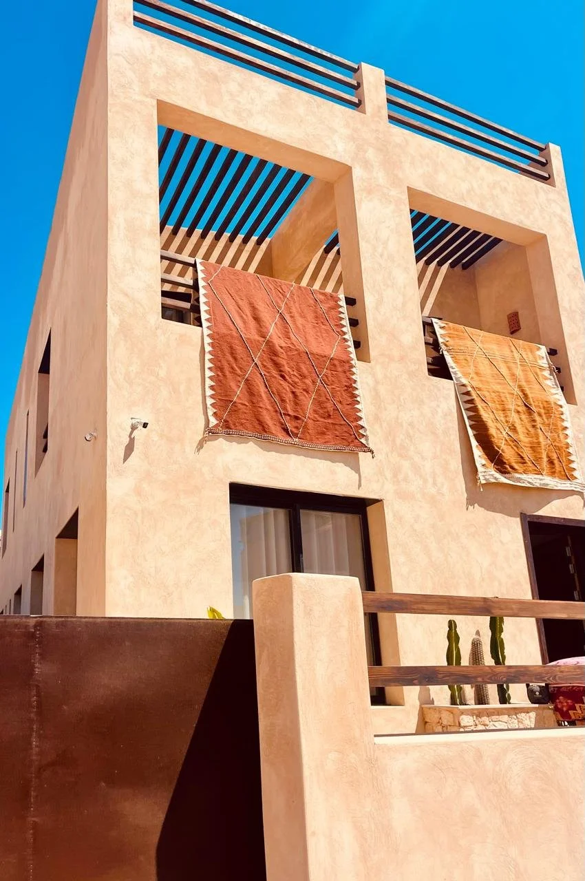 Two patterned rugs hang over a balcony of a beige stucco building with open rectangular windows, under a bright blue sky.