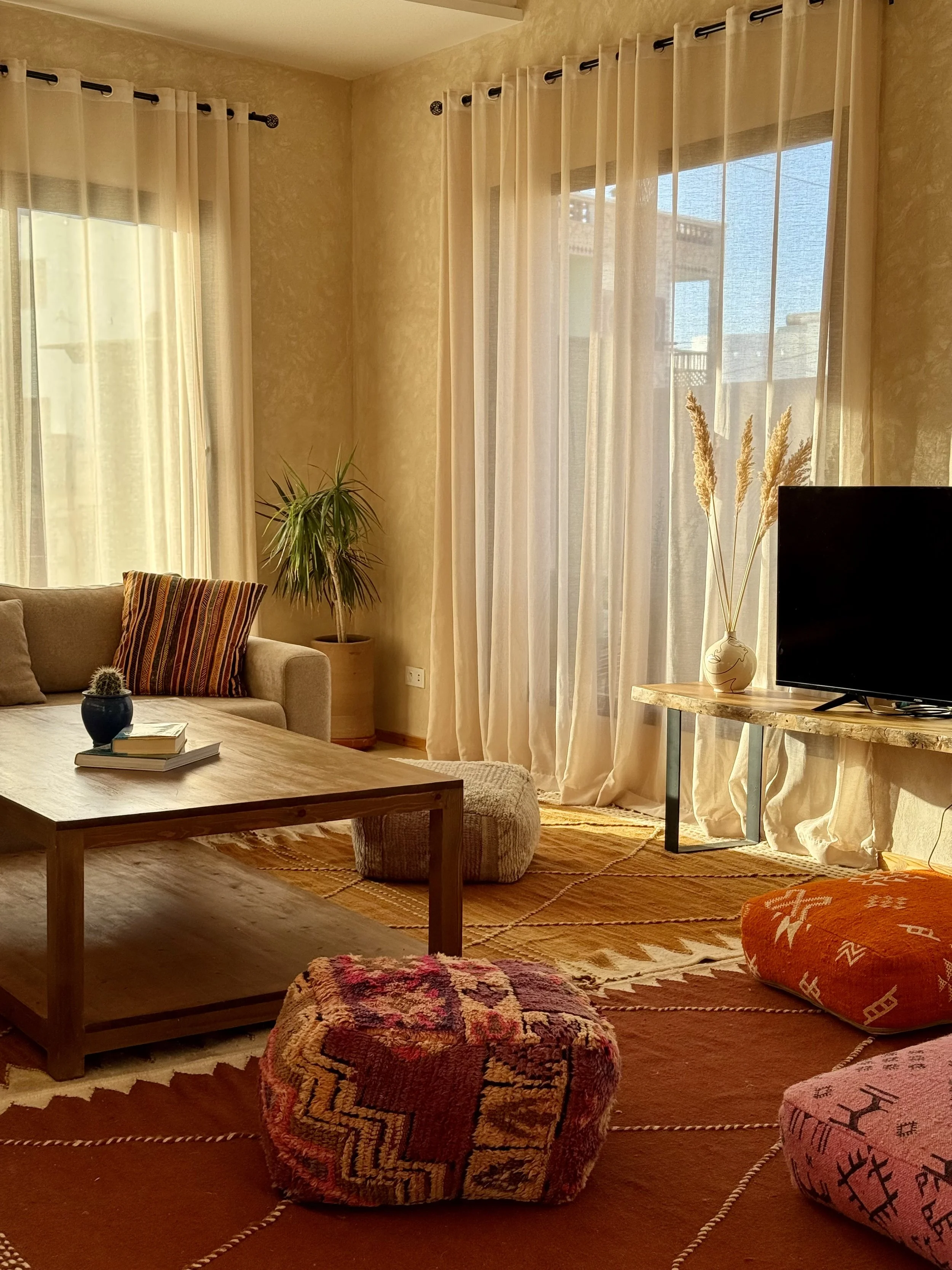 Cozy living room with beige walls, large windows with sheer curtains, a beige sofa with throw pillows, a wooden coffee table, a potted plant, a TV on a wooden stand, and floor cushions on an orange and yellow patterned rug.