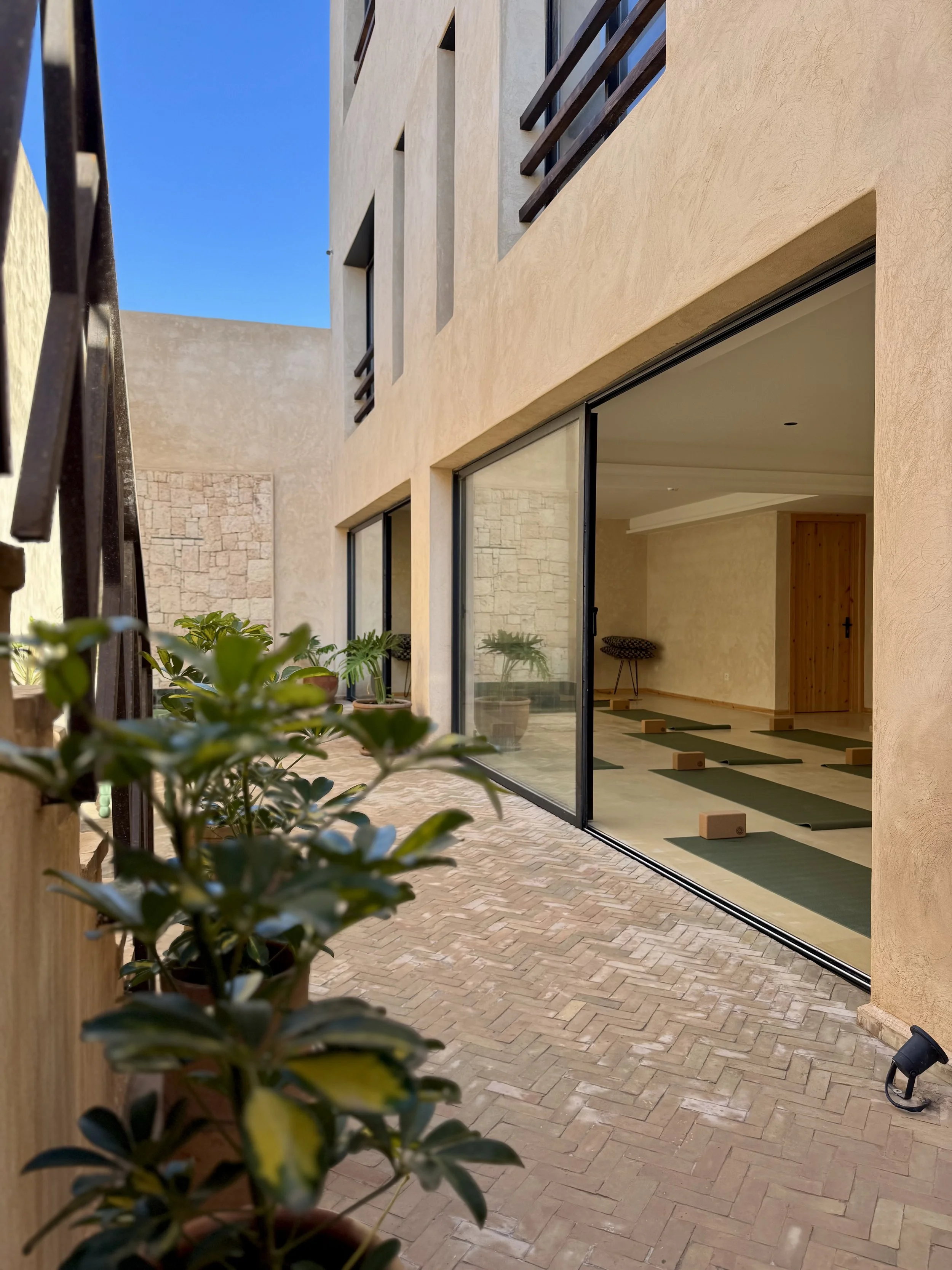 View of a courtyard with a yoga studio visible through large glass sliding doors, plants along the patio, hotel-style building with beige walls, brick flooring, and wooden accents.