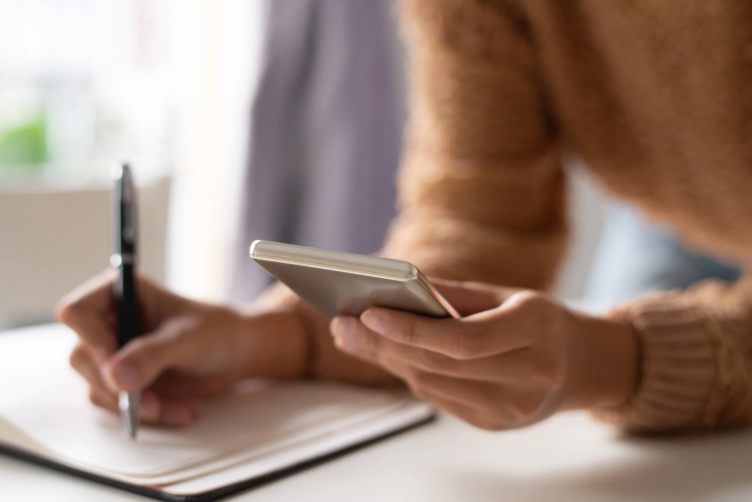Persona escribiendo en cuaderno con bolígrafo y sosteniendo teléfono móvil.
