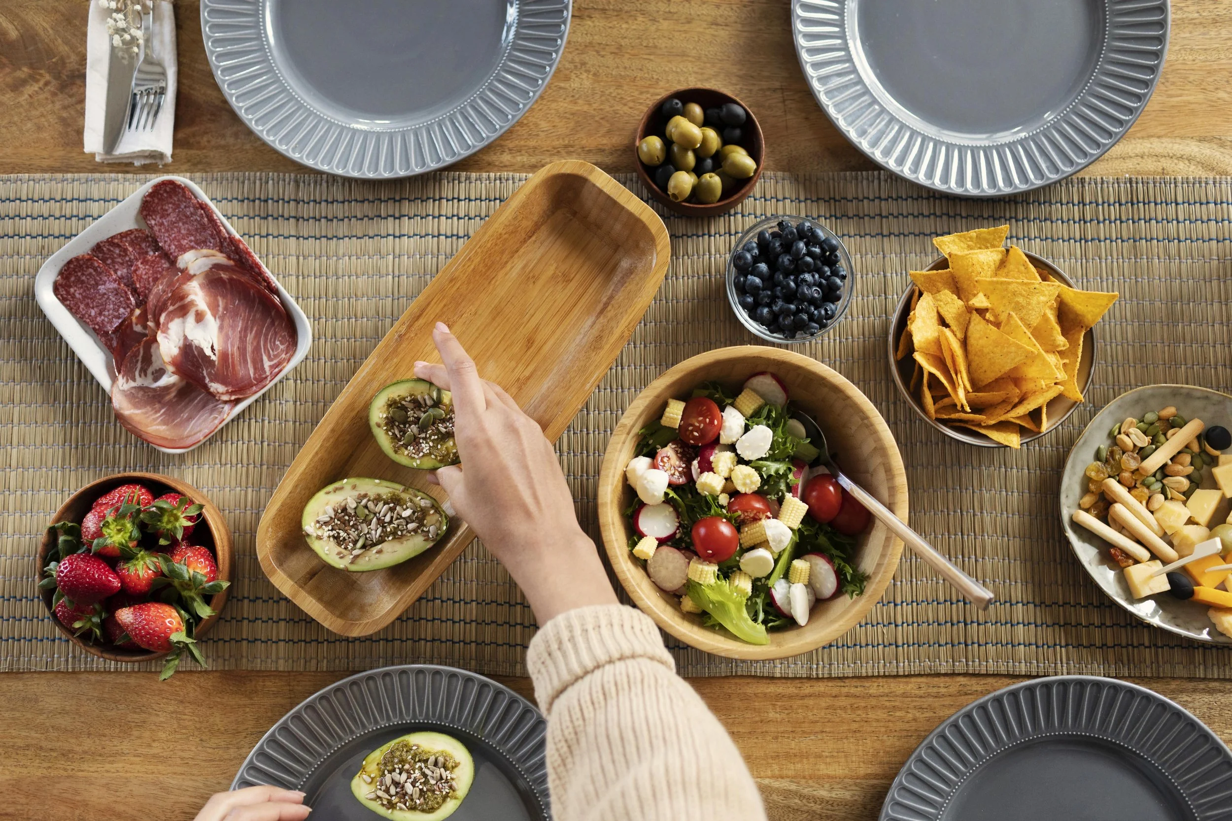 Mesa puesta con ensalada, frutos, chorizo, aguacate relleno, nopal, uvas, fresas, totopos, queso con galletas y embutidos, en platos de cerámica gris y un centro de mesa con personas sirviendo.