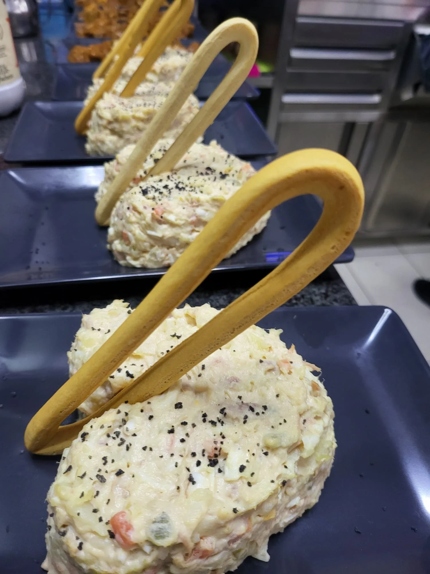 Porciones de ensalada de papa con pepinillos, huevo y especias, decoradas con galletas saladas en forma de lazo, servidas en platos negros.